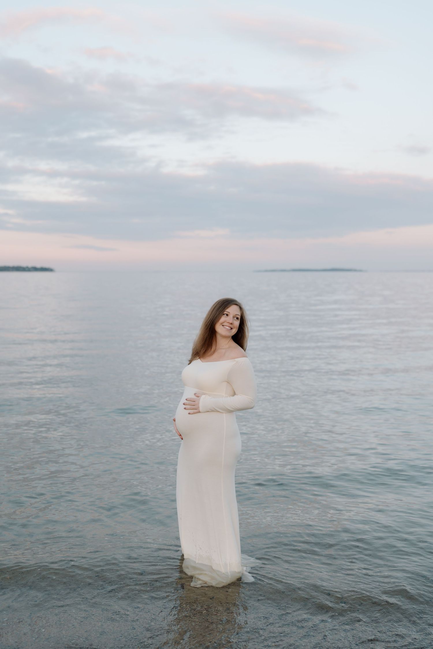 Maternity photos at sunset by calm ocean waters in flowing white dress.