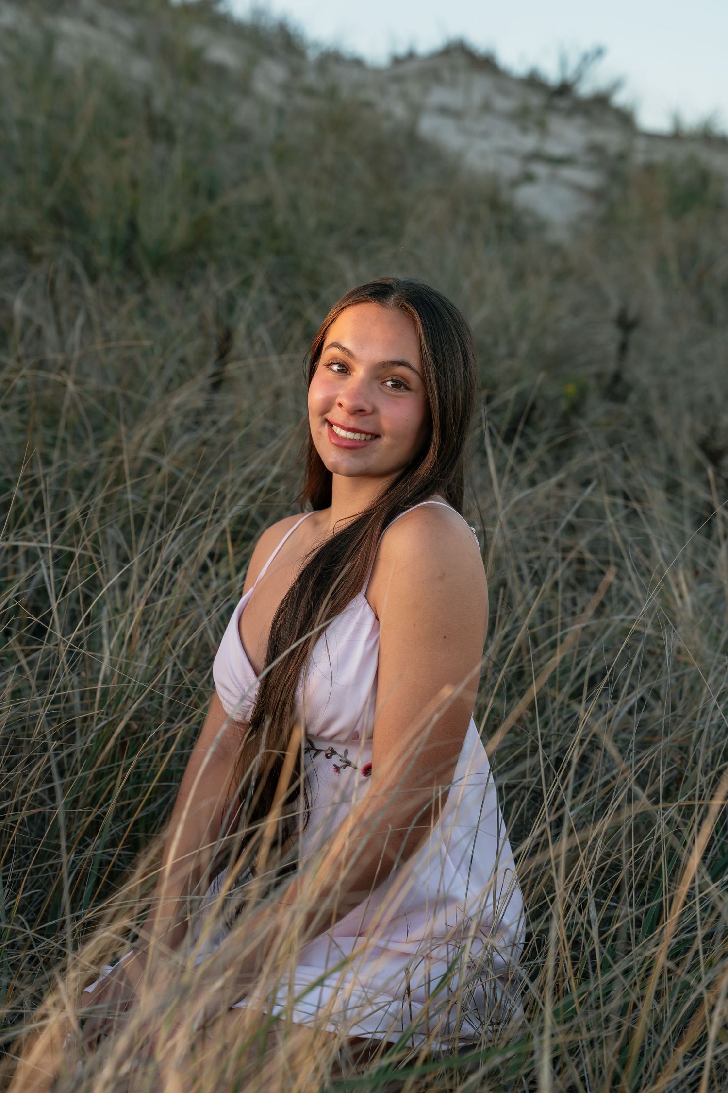 Person in white top poses among tall beach grass during sunset photoshoot.