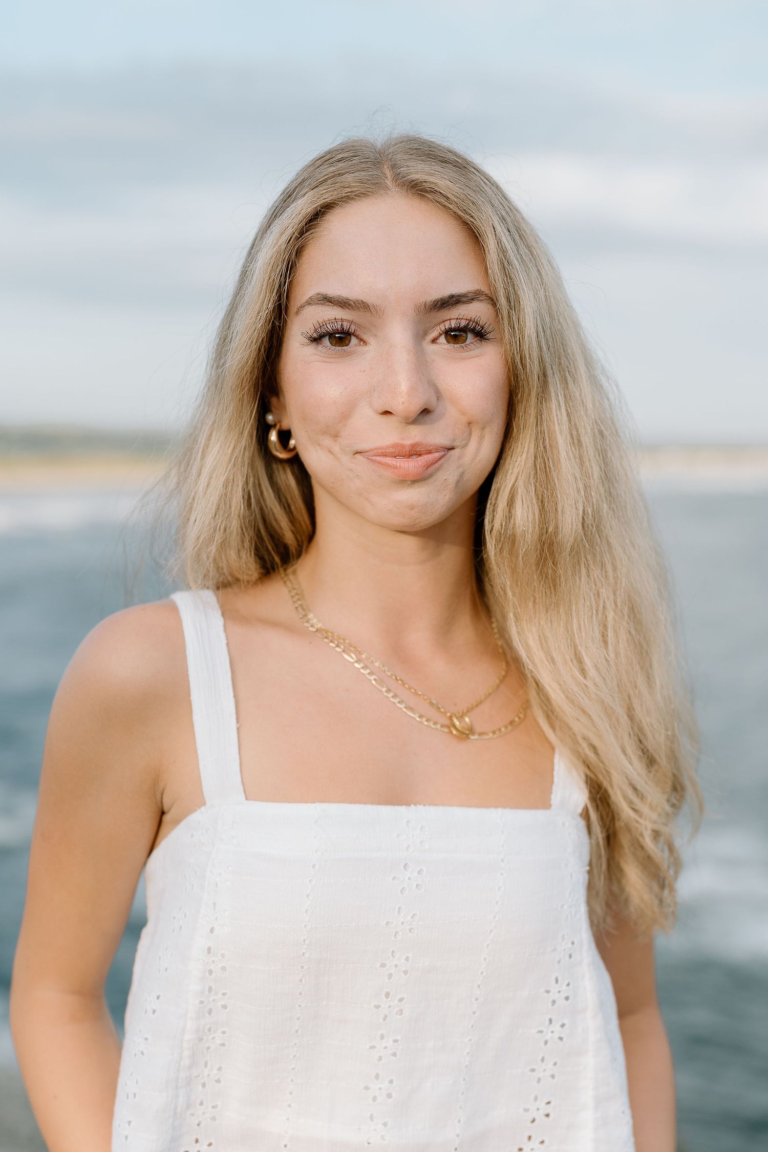 Summer portrait in white eyelet tank top with gold jewelry against ocean background.