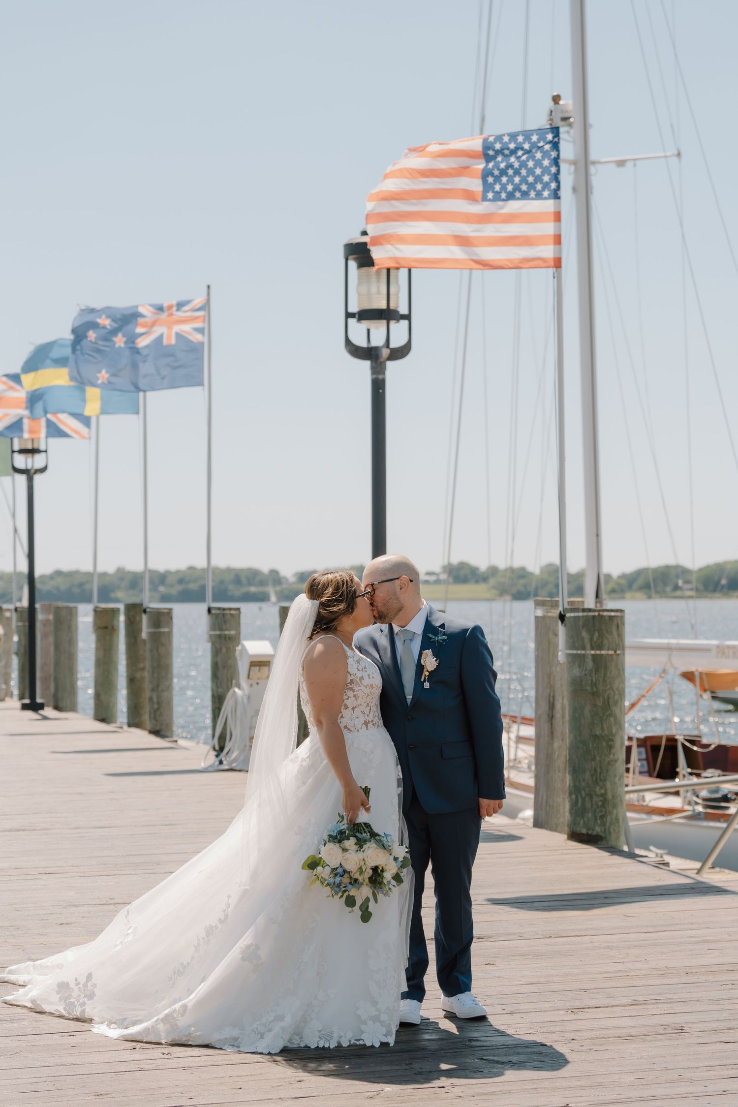 A wedding couple poses on a dock with an American flag and other flags waving in the background.