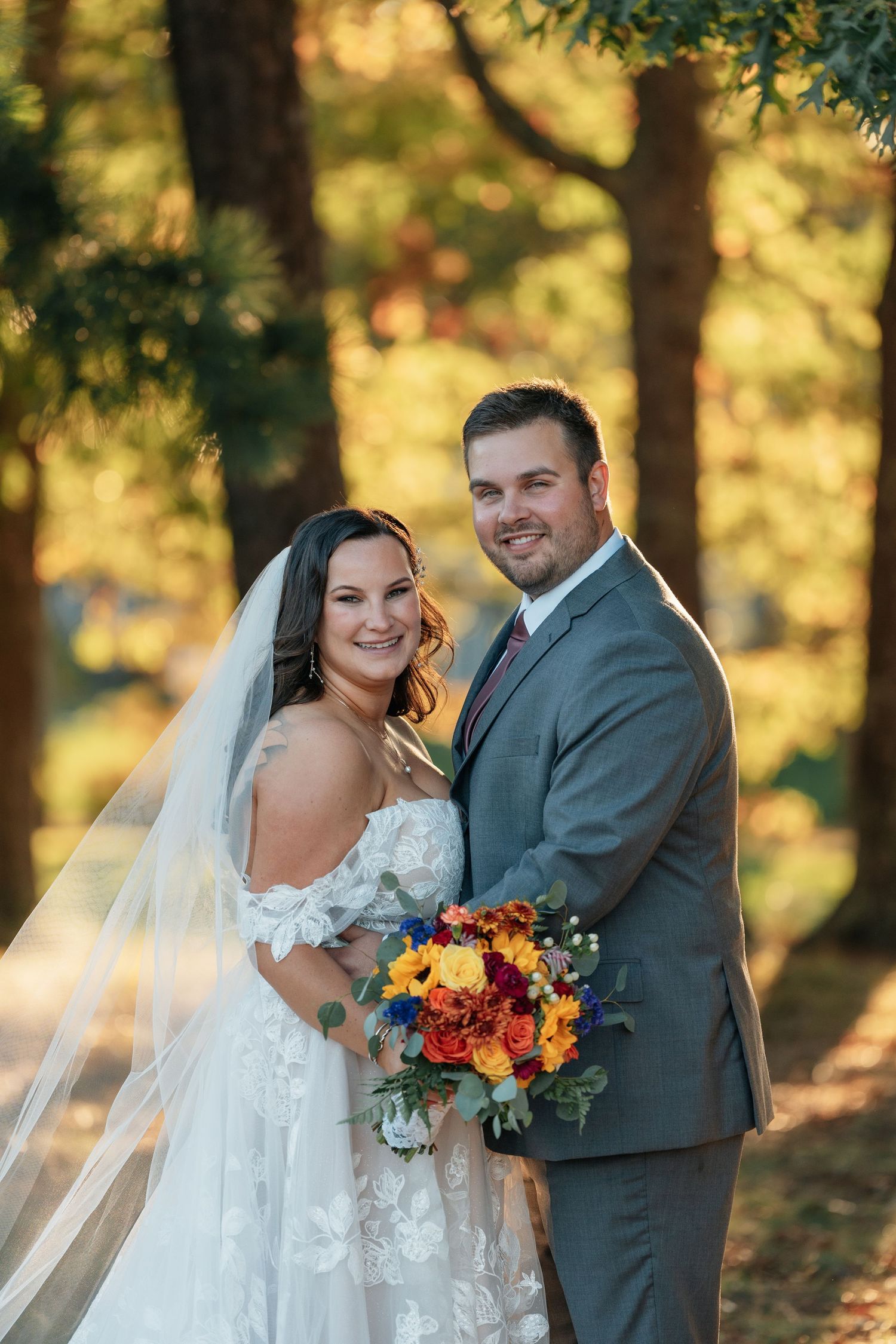 Autumn wedding photoshoot featuring a couple with bright orange and yellow bouquet among trees.