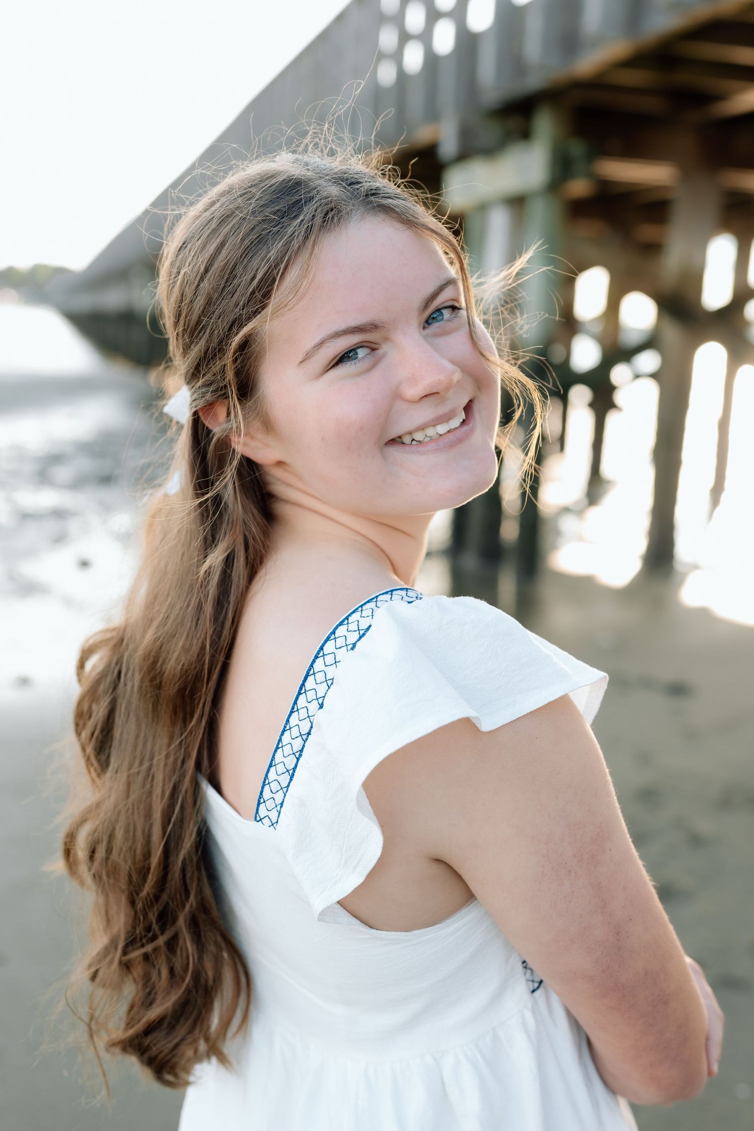 Beach portrait under pier featuring someone in white dress with embroidered details.