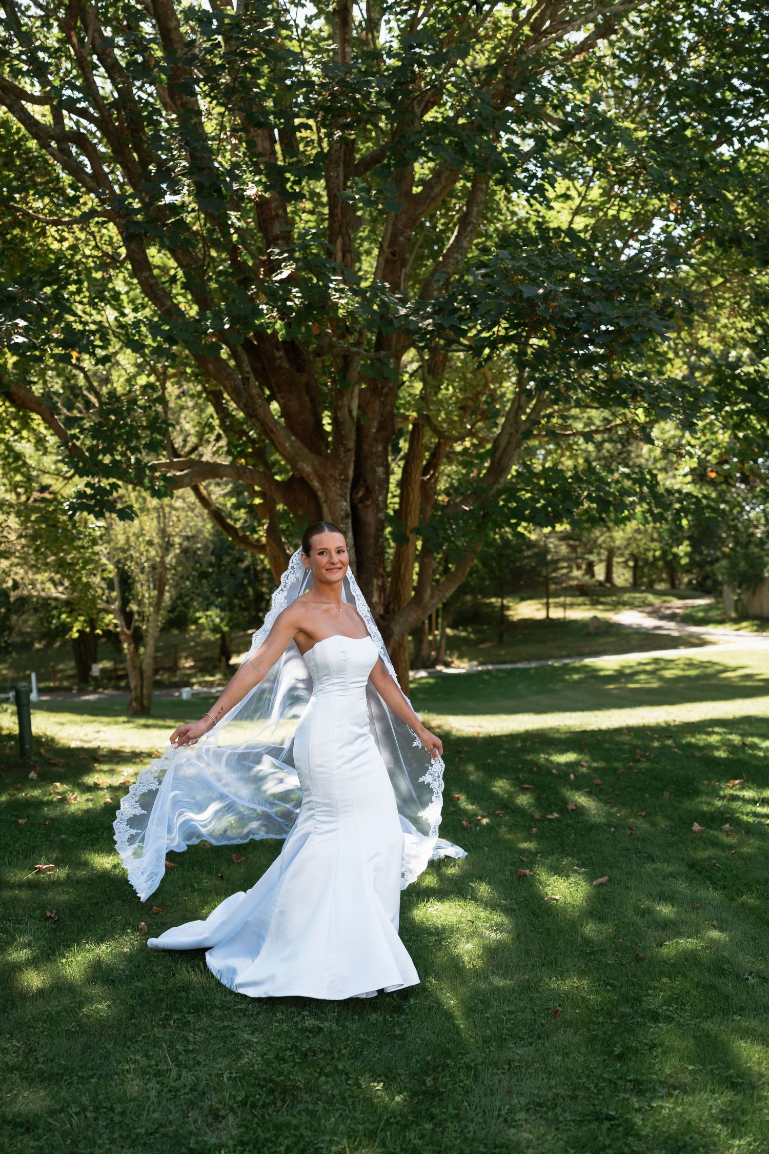 White wedding dress flowing in the breeze on a sunny lawn with trees in the background.