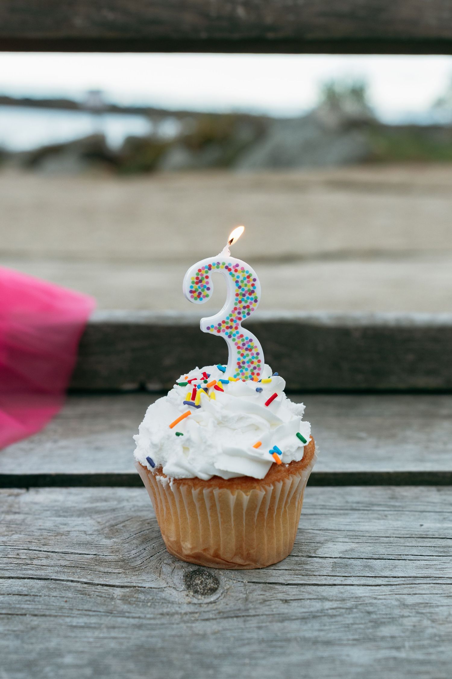 Birthday cupcake with white frosting and sprinkles topped with number 3 candle on wooden surface.
