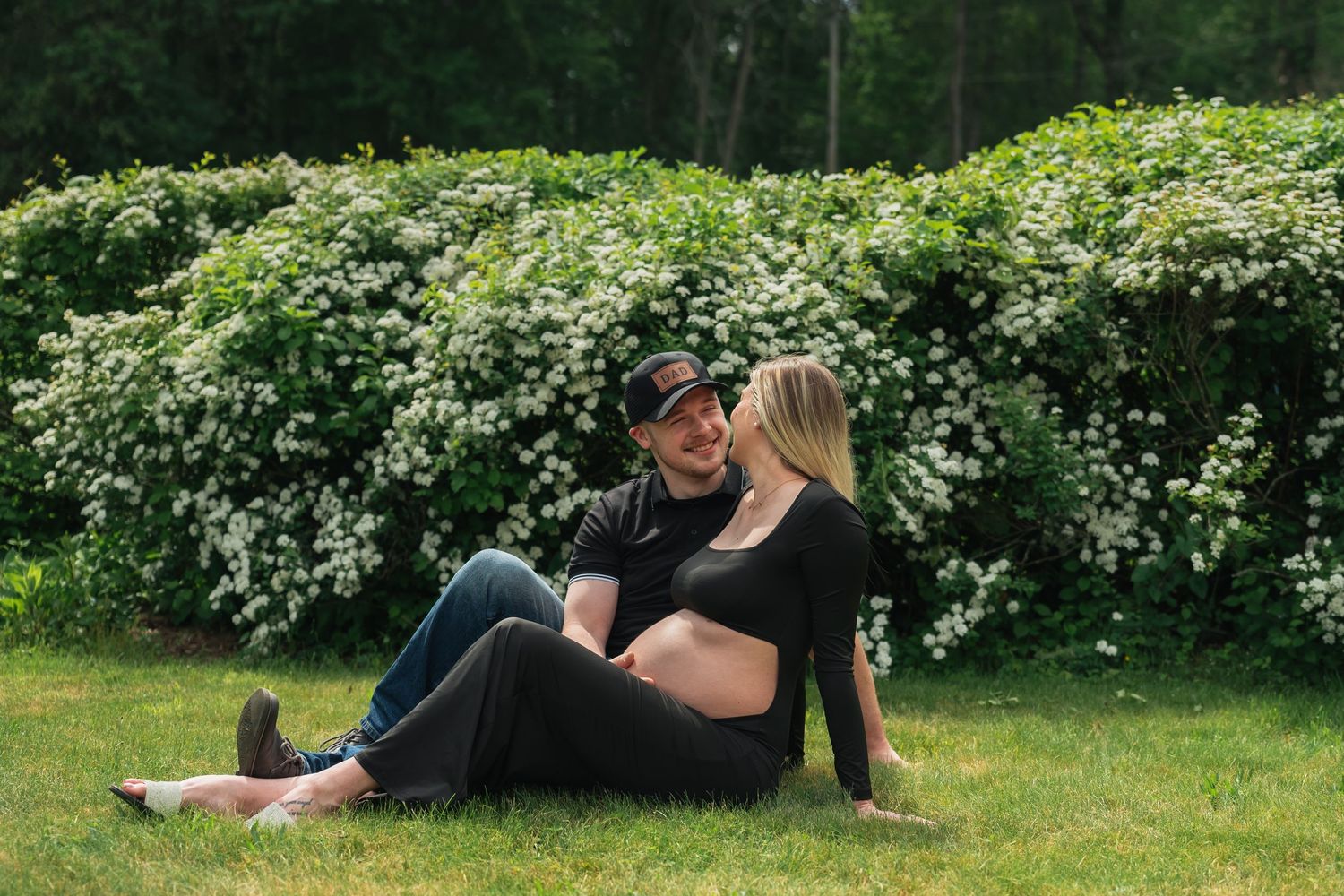 A couple sits together on grass near flowering bushes in a garden setting.