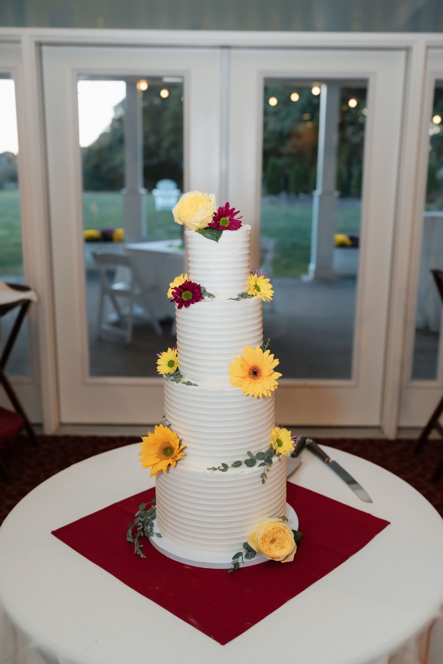 Four-tier white wedding cake decorated with yellow and burgundy flowers on a red napkin.