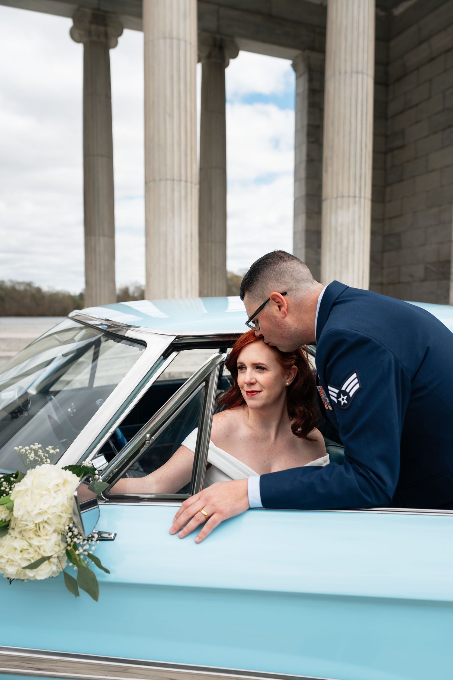 Wedding couple posing with vintage light blue car near classical columns.