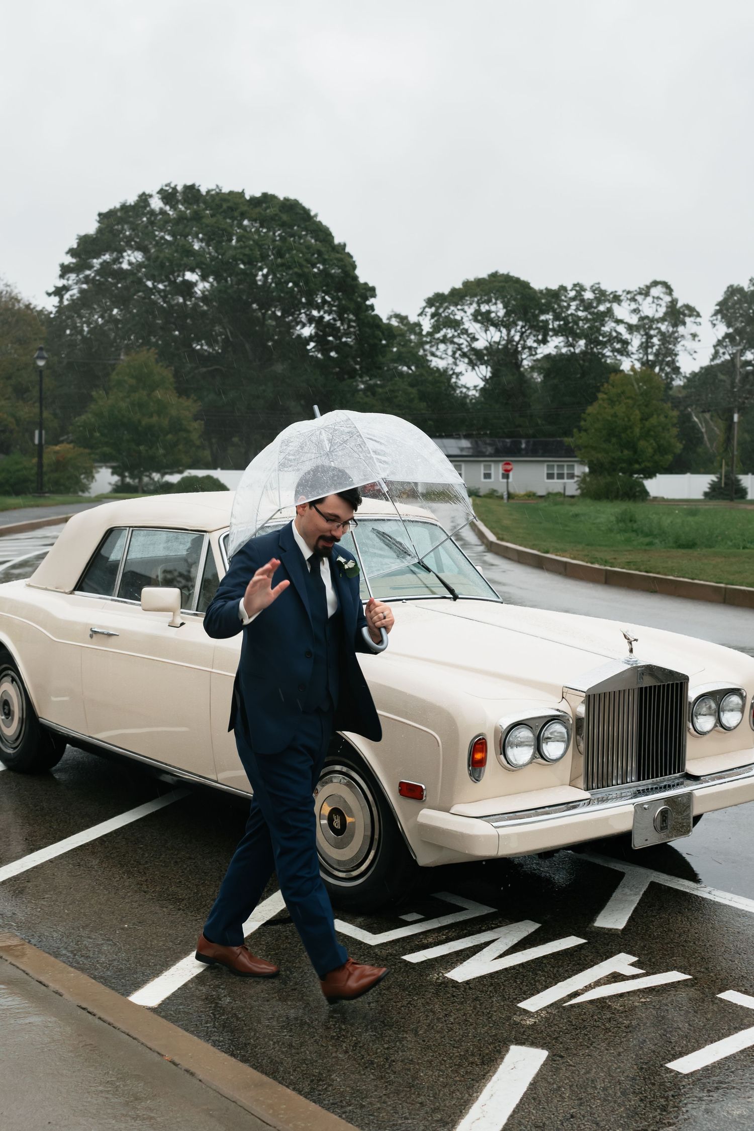 Groom with clear umbrella walks past classic white Rolls Royce in rainy parking lot.