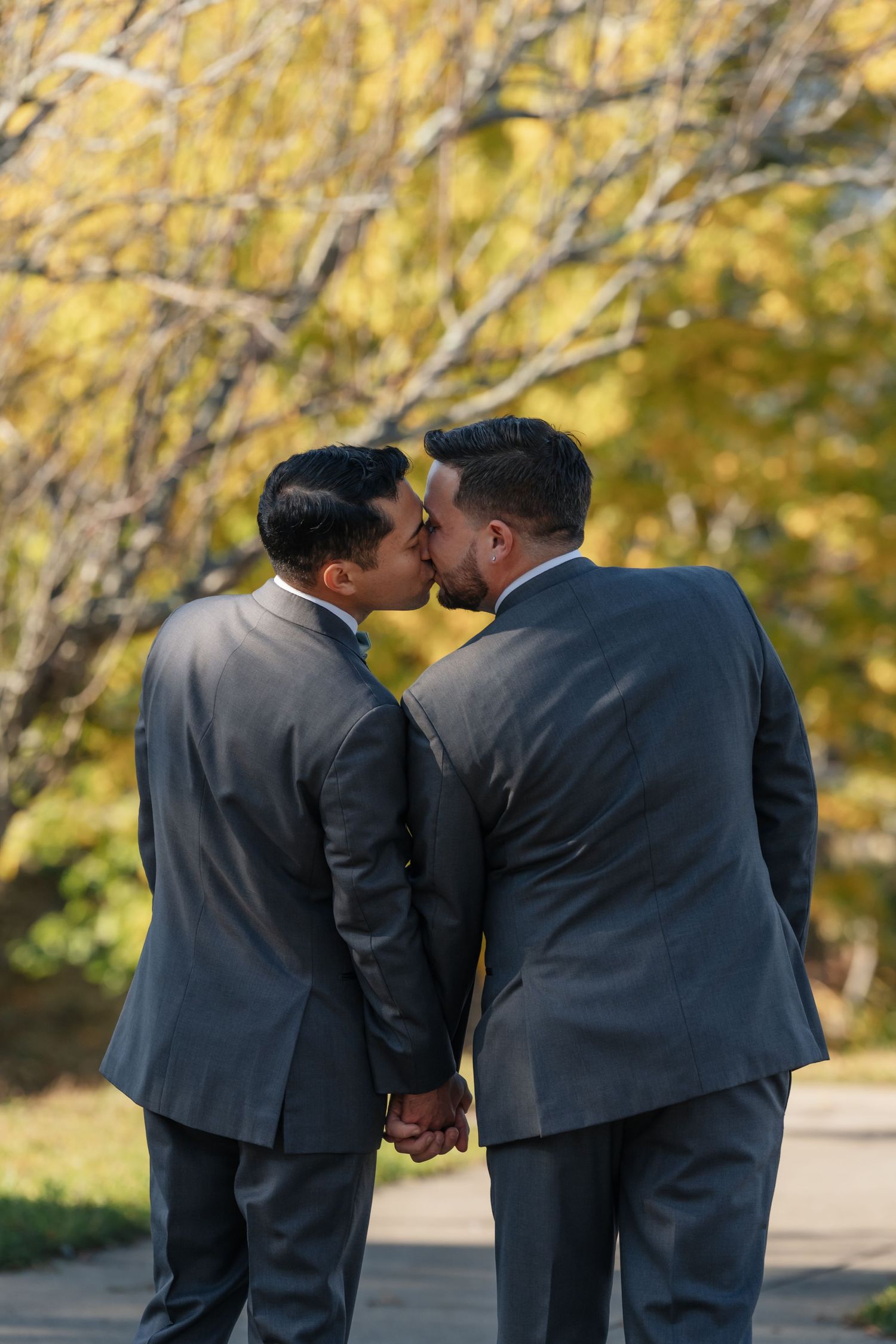 Two people in gray suits share a romantic moment against autumn trees.