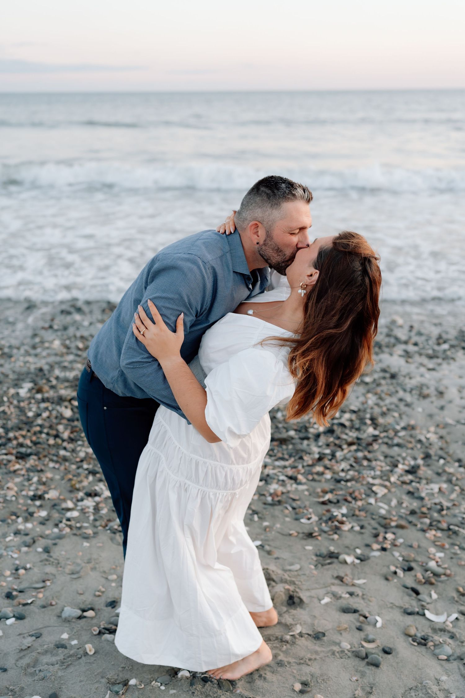 Romantic couple embraces on a rocky beach at sunset, with waves crashing in the background.