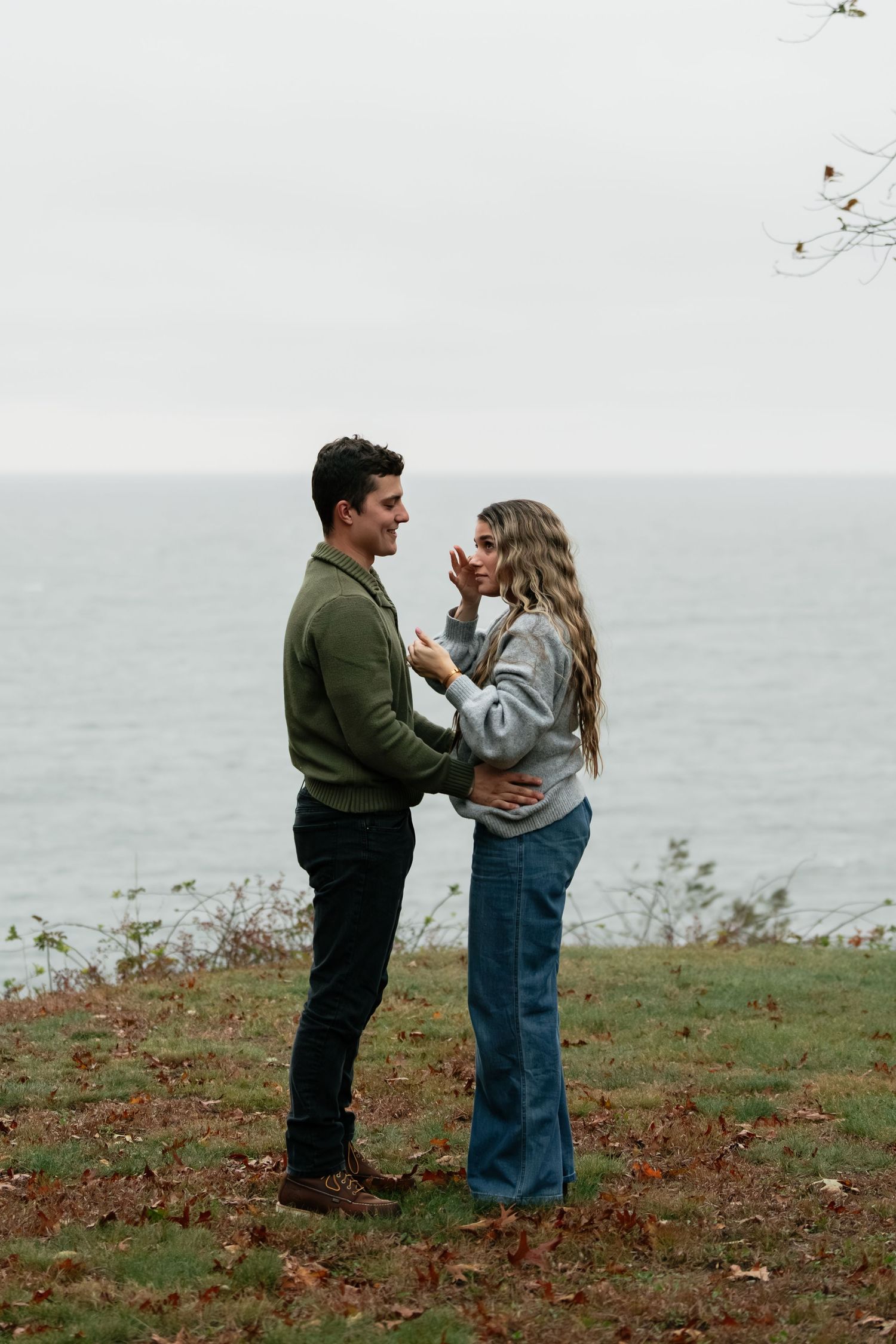 A couple stands together on an autumn hillside overlooking the sea.