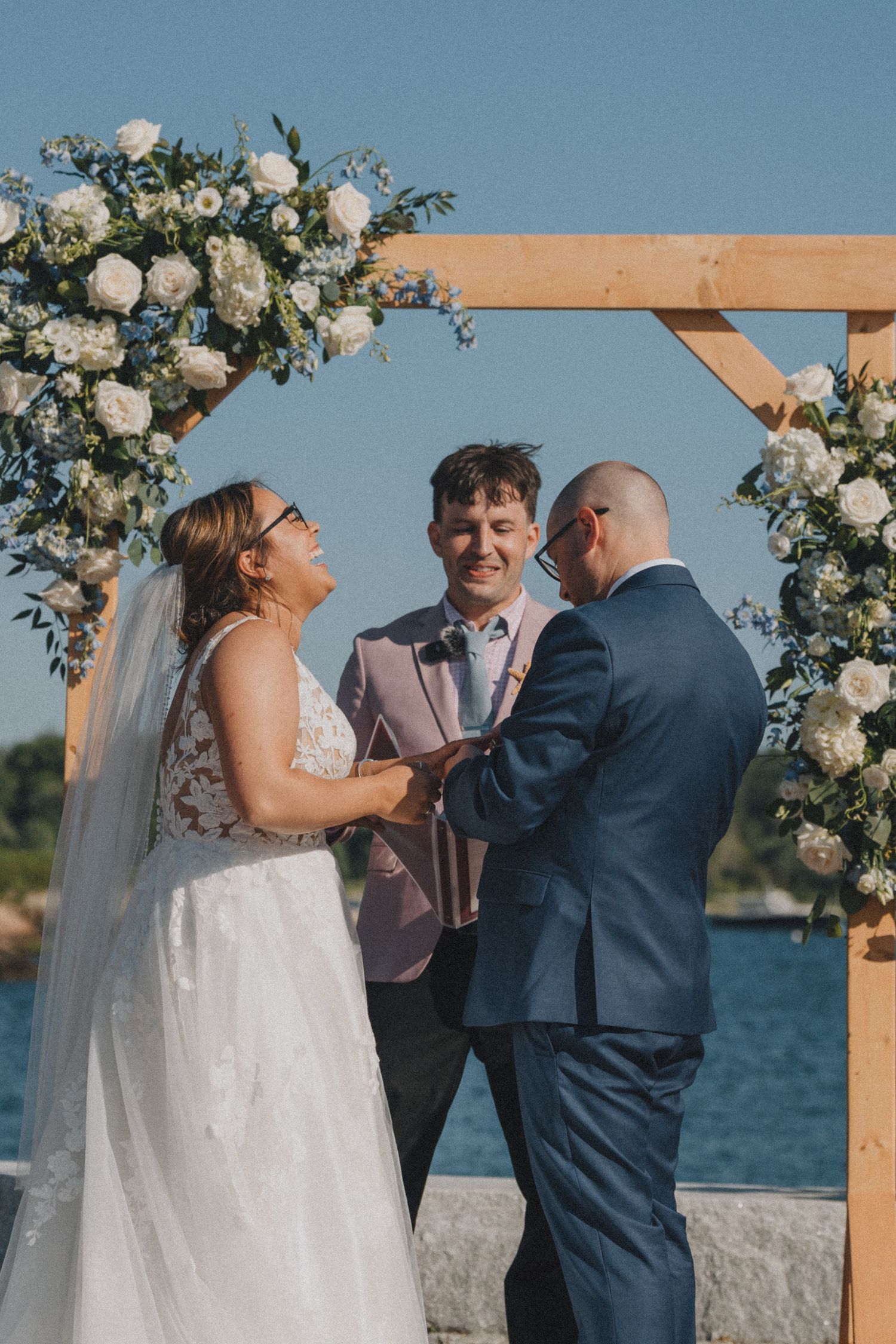 Waterfront wedding ceremony under a floral arch with white roses and greenery.