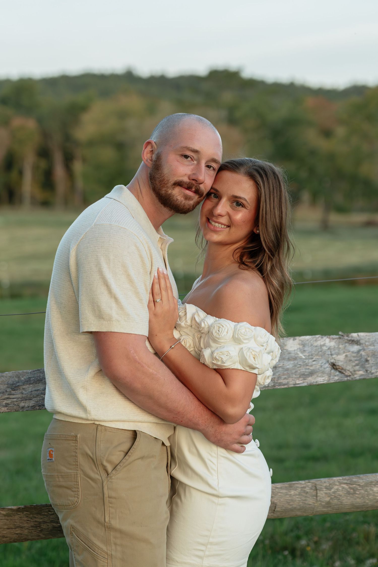 A couple embraces by a rustic fence during a countryside photoshoot at sunset.