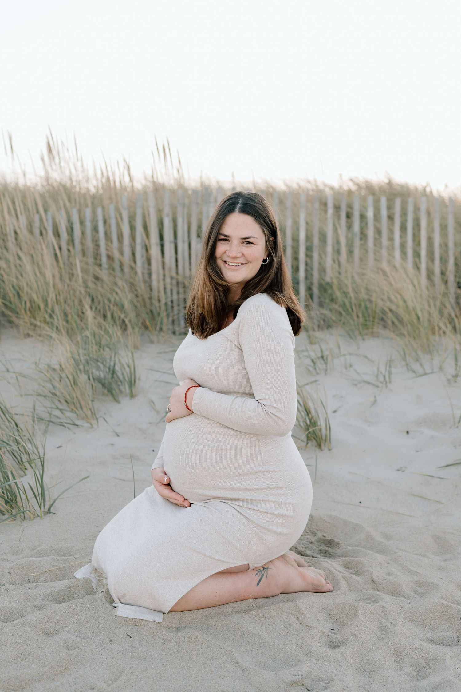 A woman in a cream dress poses on sandy beach dunes during a maternity photoshoot.