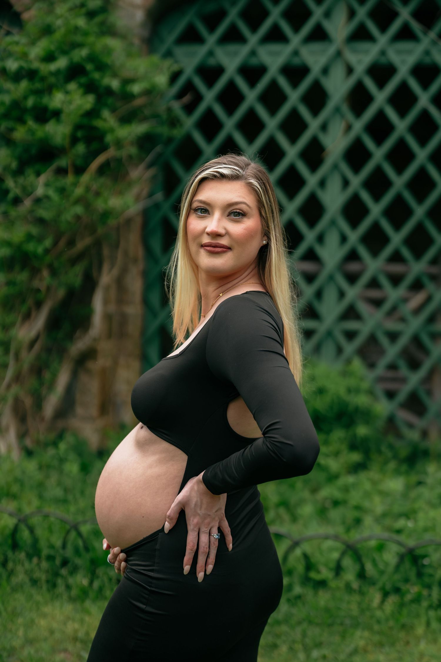 An expectant mother in a black dress poses gracefully against a garden trellis background.