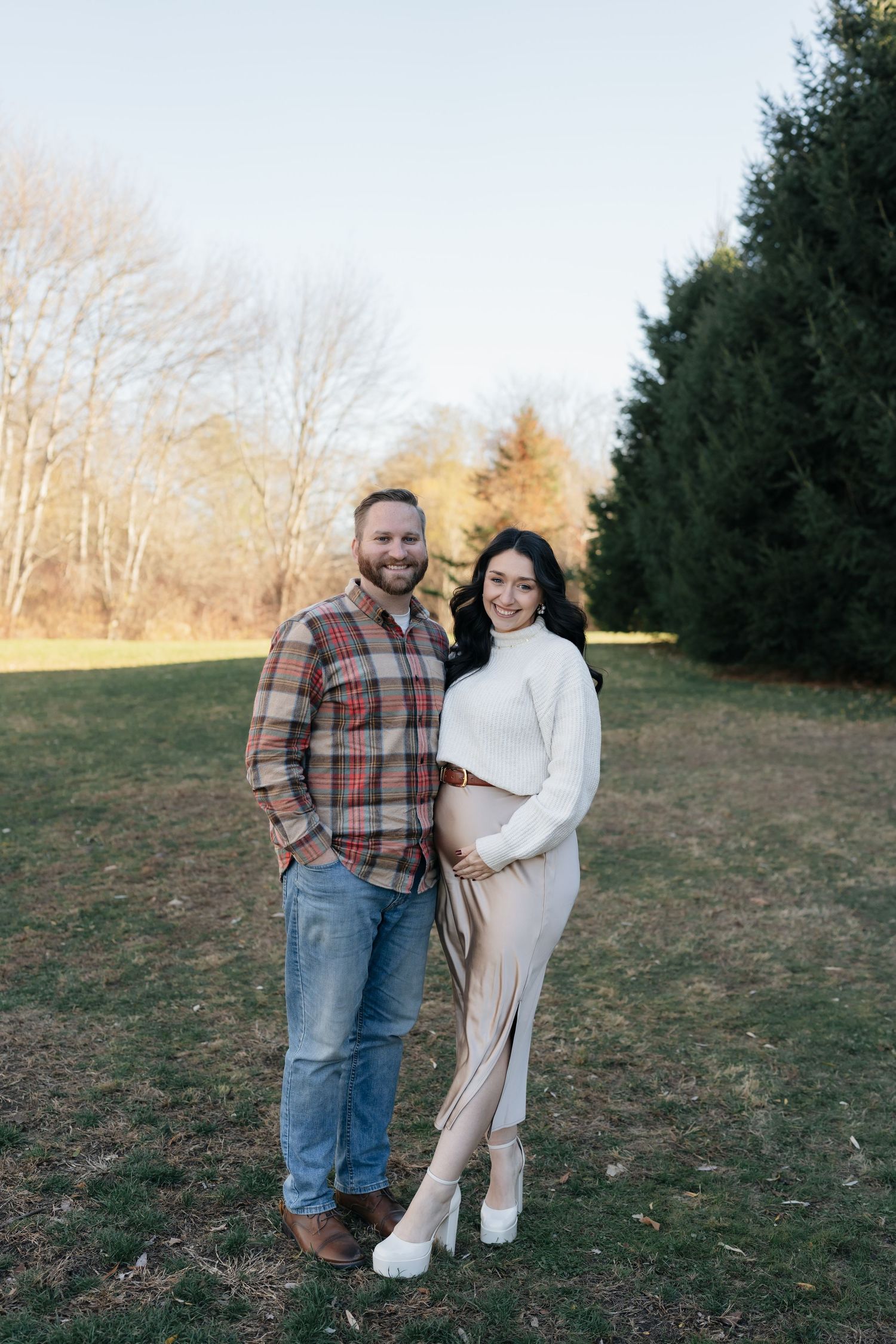 Expectant couple embraces while standing in a field during golden hour.