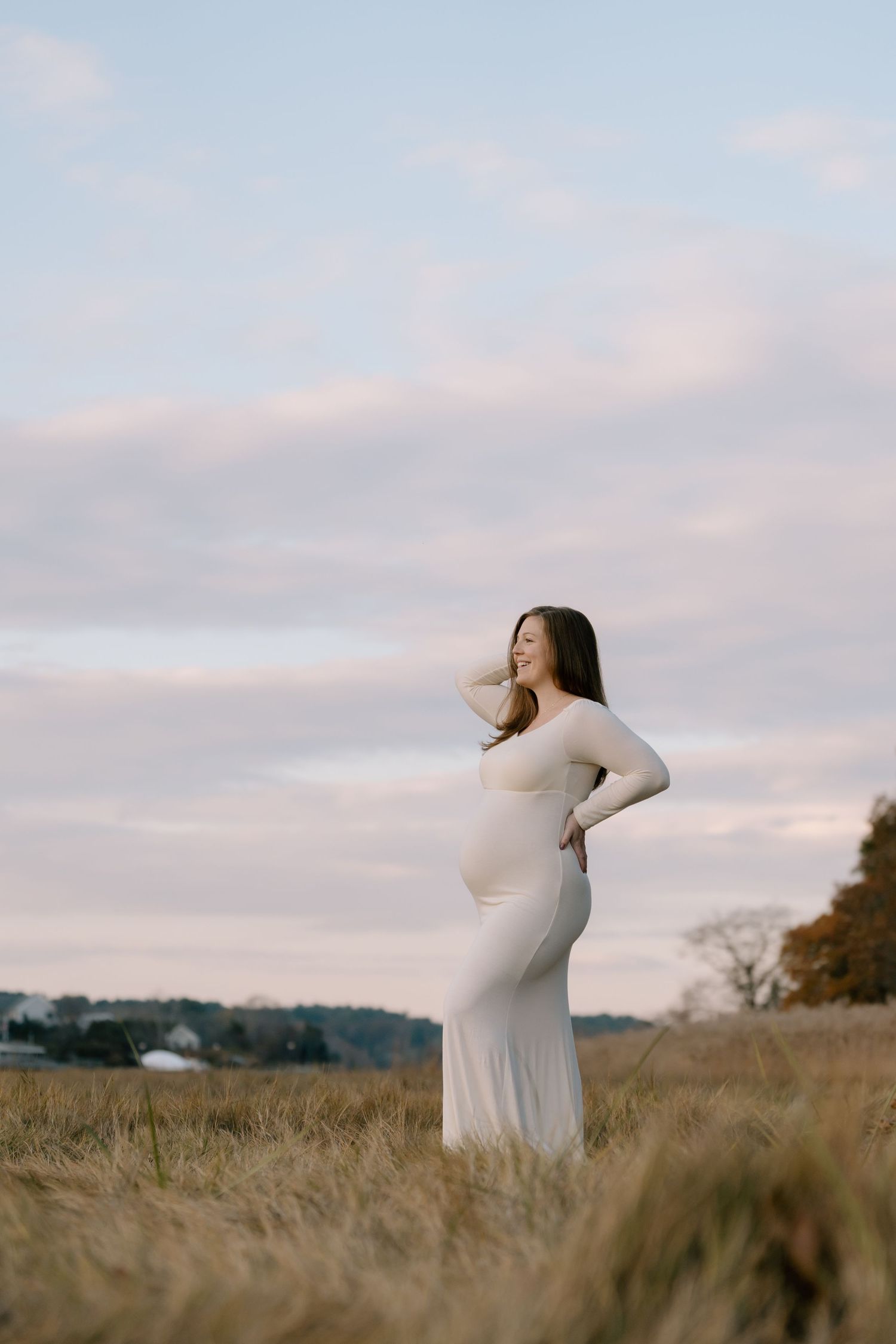 A woman in a white dress poses in a field during golden hour for a maternity photo.