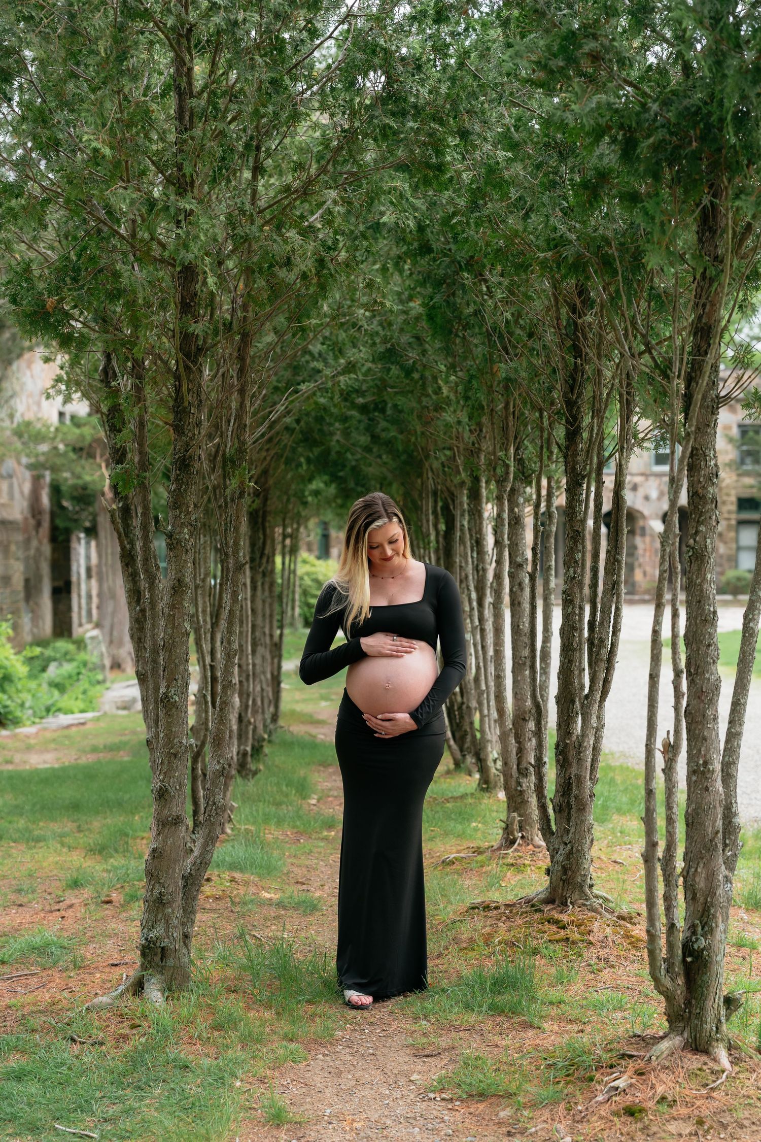A maternity photo series captured in a wooded area with tall trees creating a natural pathway.