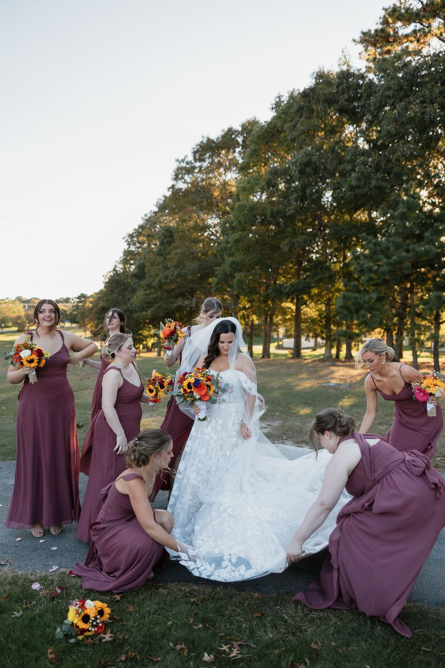 Bridesmaids in purple dresses pose playfully on grass with the bride in a white gown.