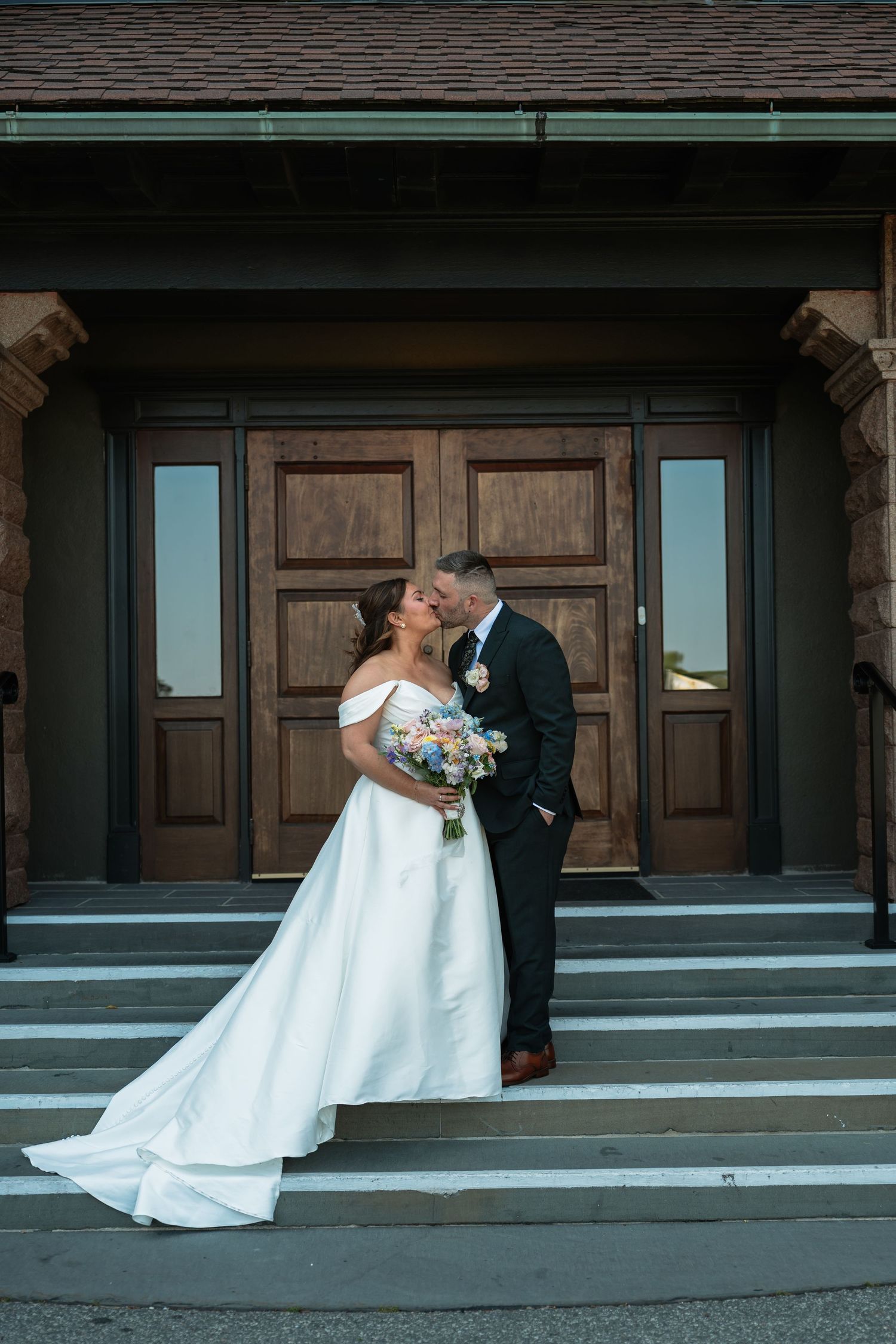 Newlyweds share a romantic moment on church steps in front of wooden doors.