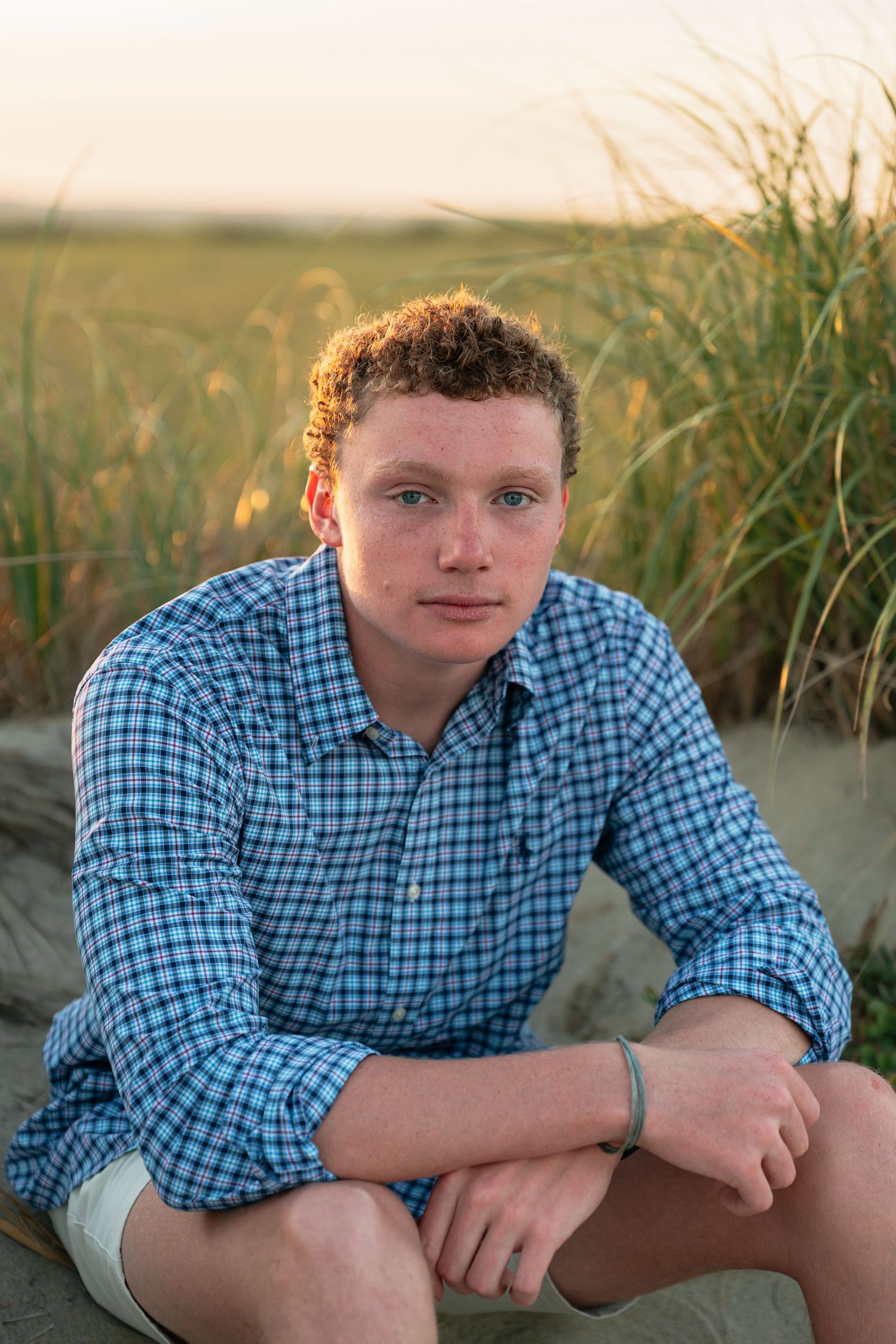Person in blue plaid shirt sits among beach grass during sunset photo shoot.