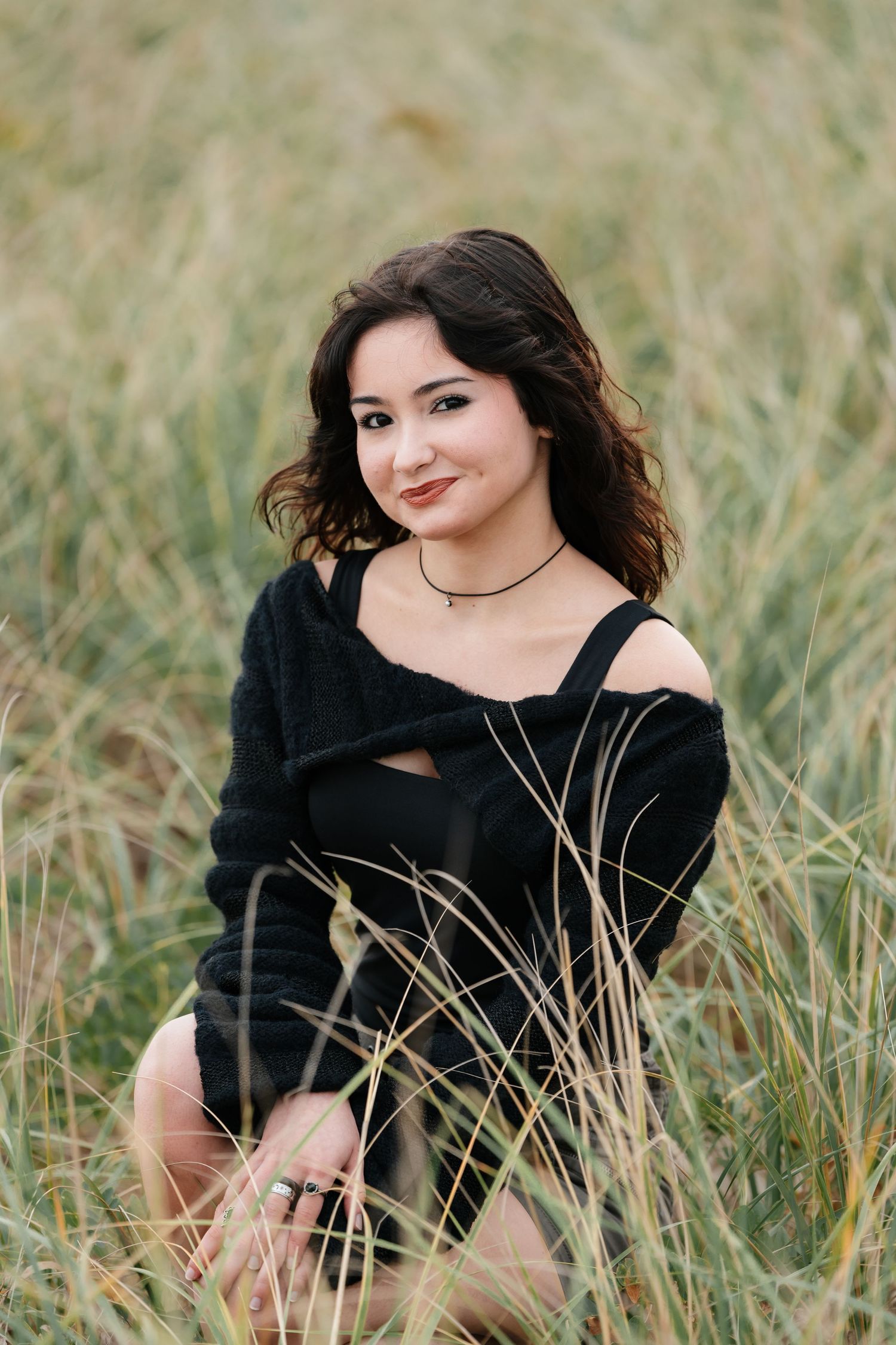 Person in black top sitting among tall beach grass during photoshoot.