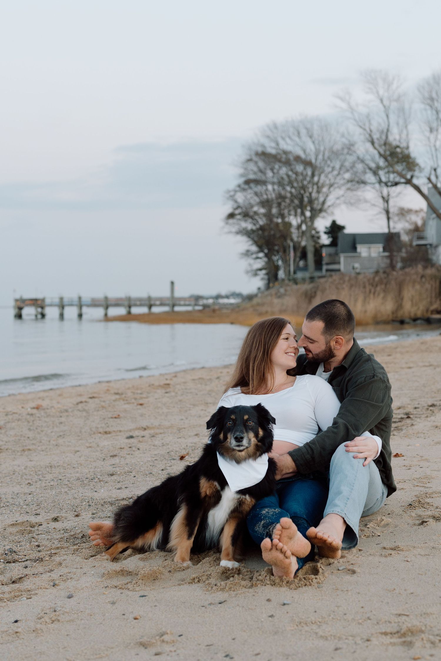 A couple sits on a beach with their black and brown dog near a wooden pier at sunset.