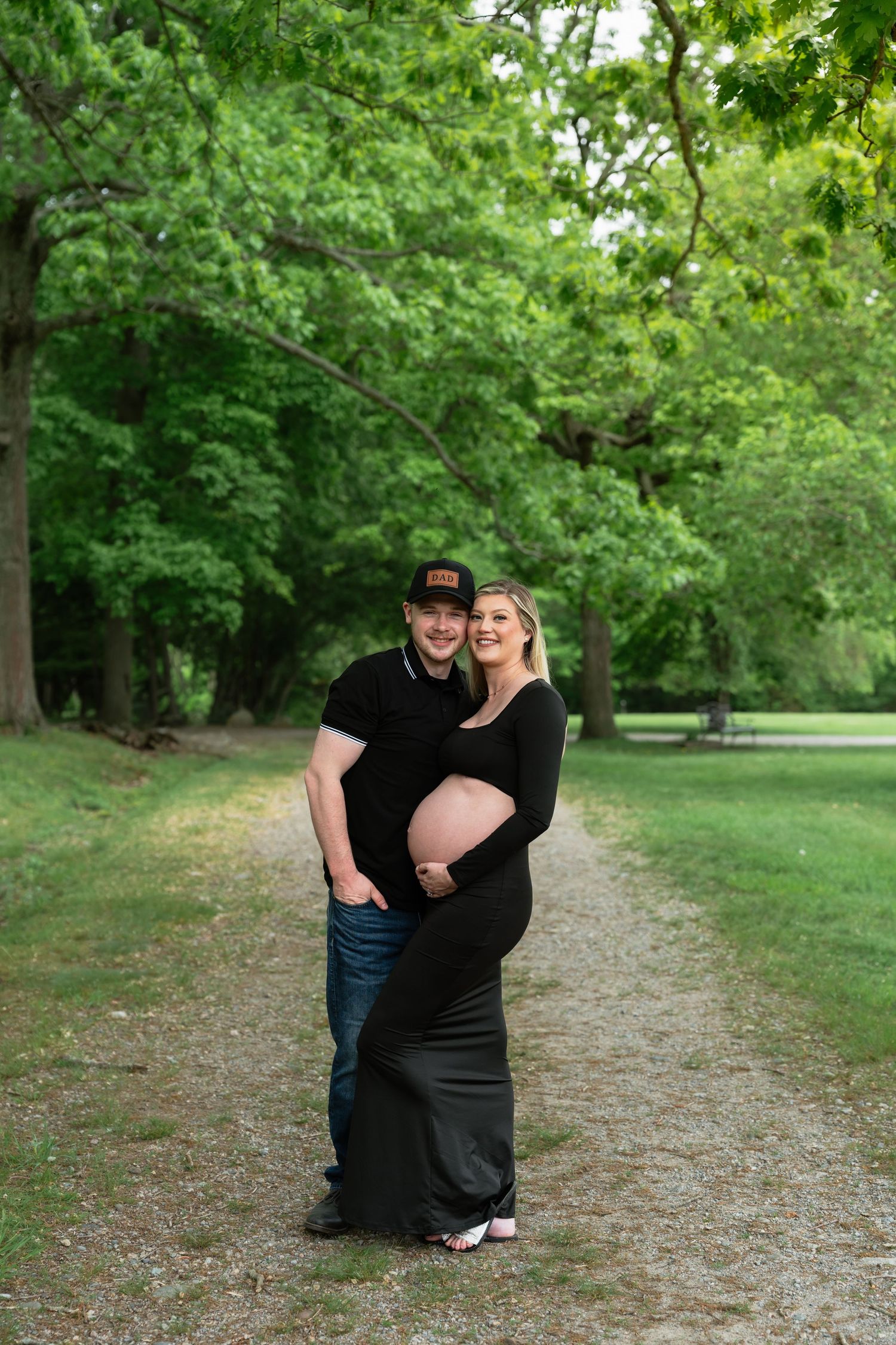 Romantic maternity photos on tree-lined path with couple in black attire.