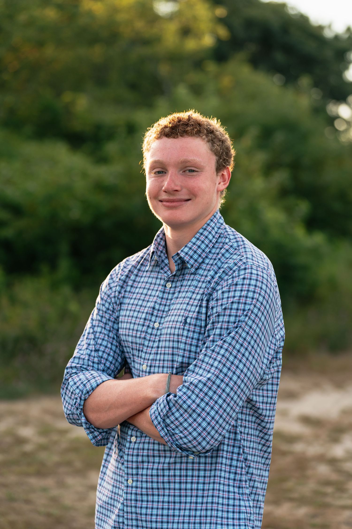 Person wearing blue checkered shirt poses outdoors with crossed arms during photo session.
