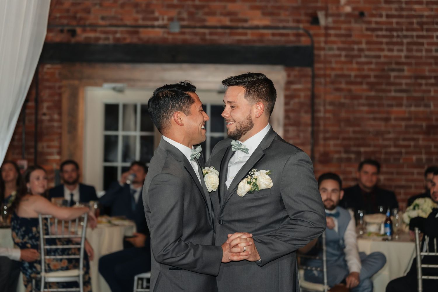 Wedding guests dancing and celebrating on a rustic brick-walled dance floor at an indoor reception venue.