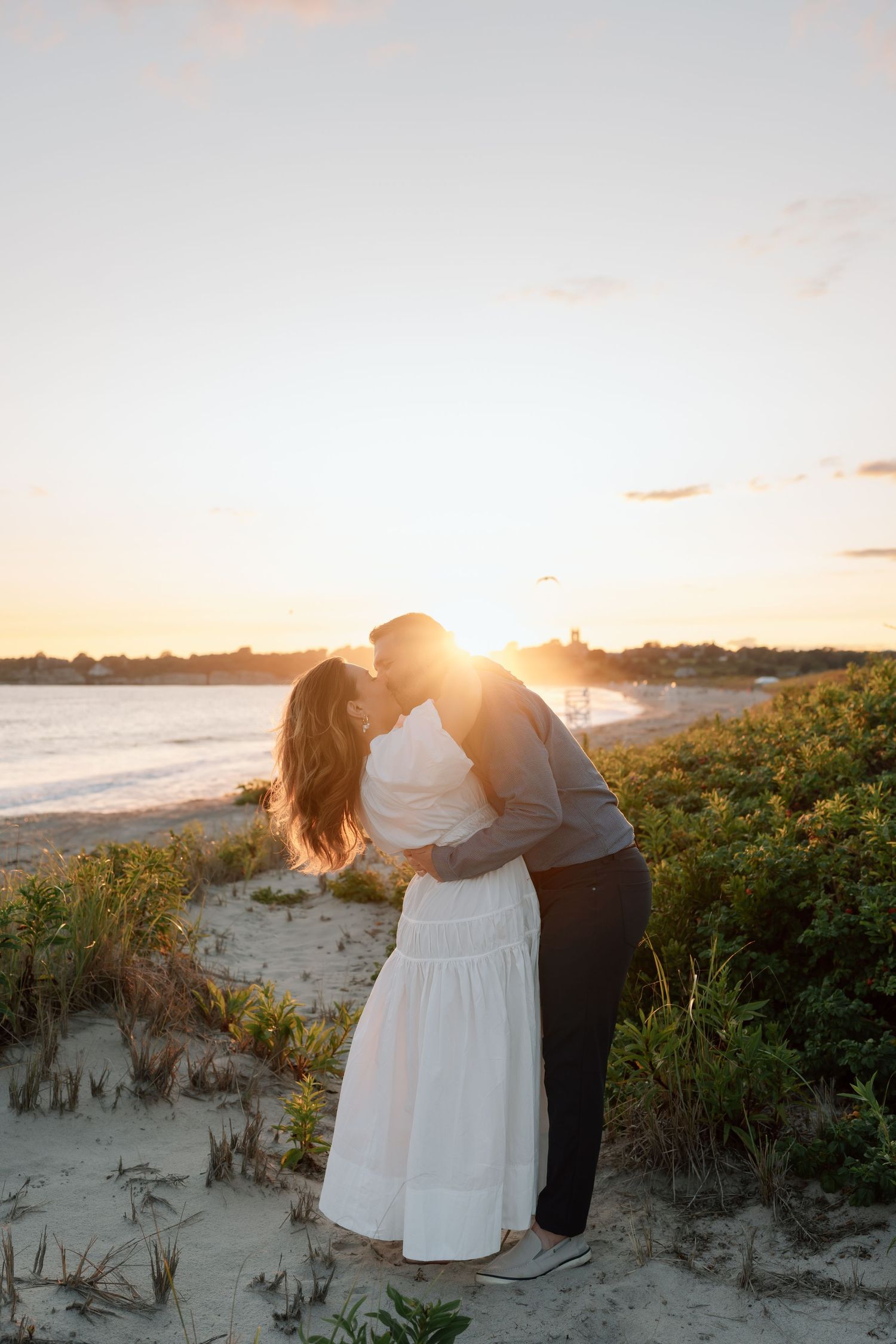 Couple shares tender moments on a beach at sunset with sand dunes and coastal vegetation.