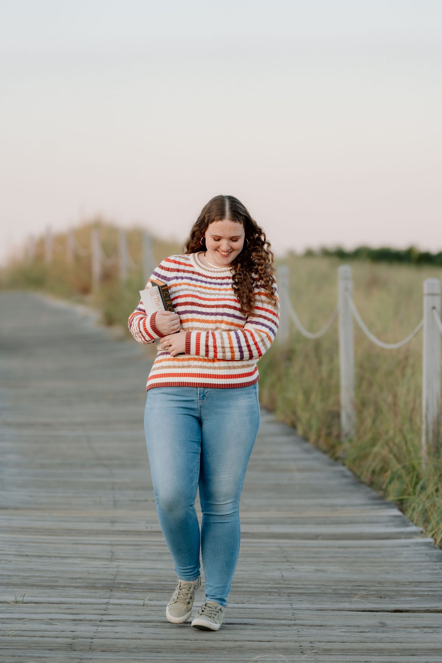 Person in striped sweater walking along wooden boardwalk path surrounded by coastal grass.