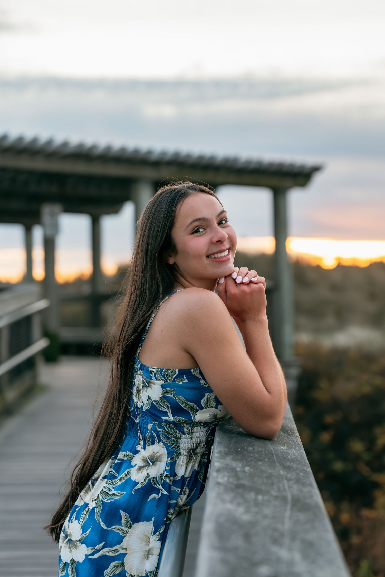 Person in blue floral dress posing by wooden boardwalk at sunset.