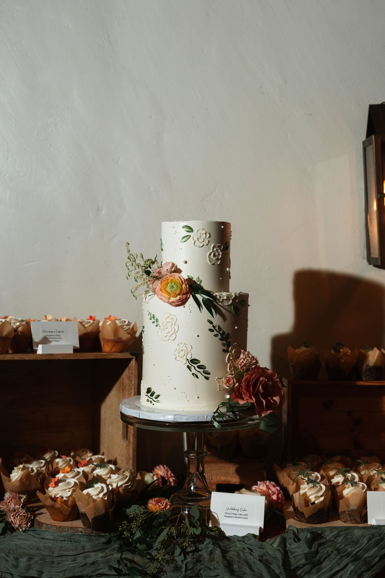 White tiered wedding cake decorated with floral designs displayed alongside dessert table.