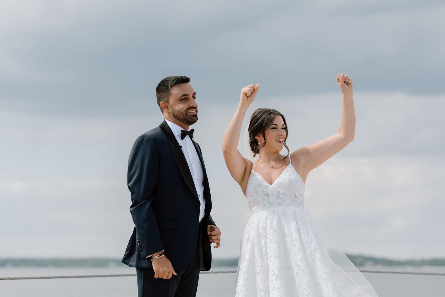 Joyful wedding couple celebrating by the ocean with dramatic cloudy backdrop.