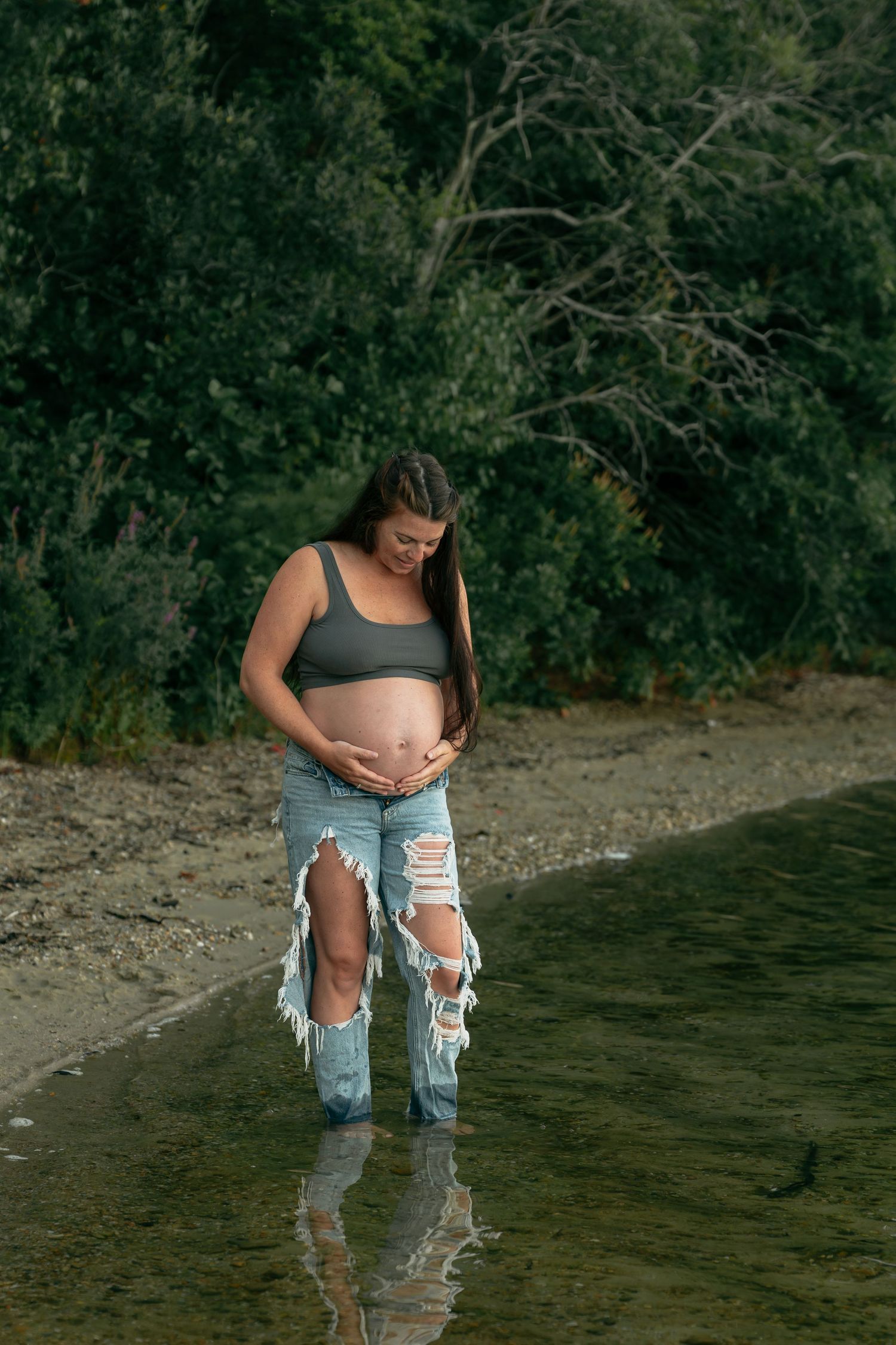 Expectant mother in distressed jeans stands in shallow water at lakeside during maternity photoshoot.