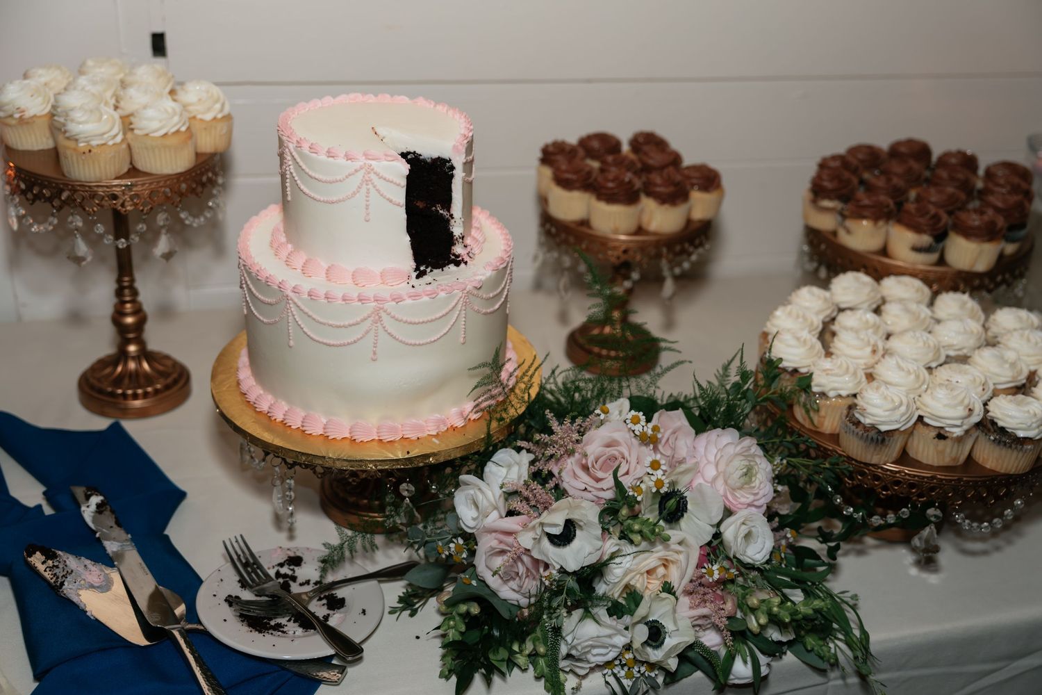 Wedding dessert table with two-tier pink cake, cupcakes and floral decorations.