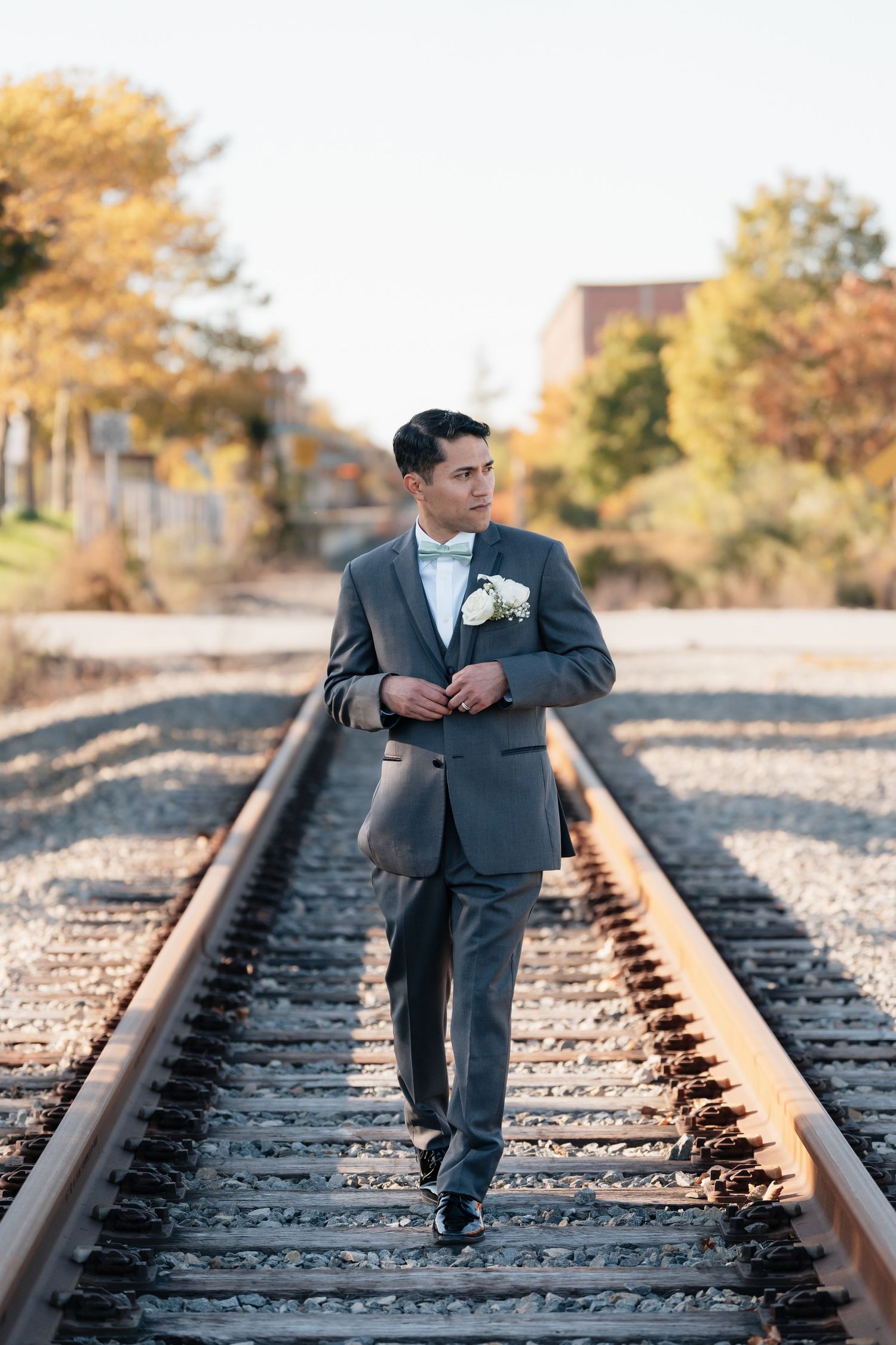 A groom in a gray suit poses on railroad tracks during golden hour.