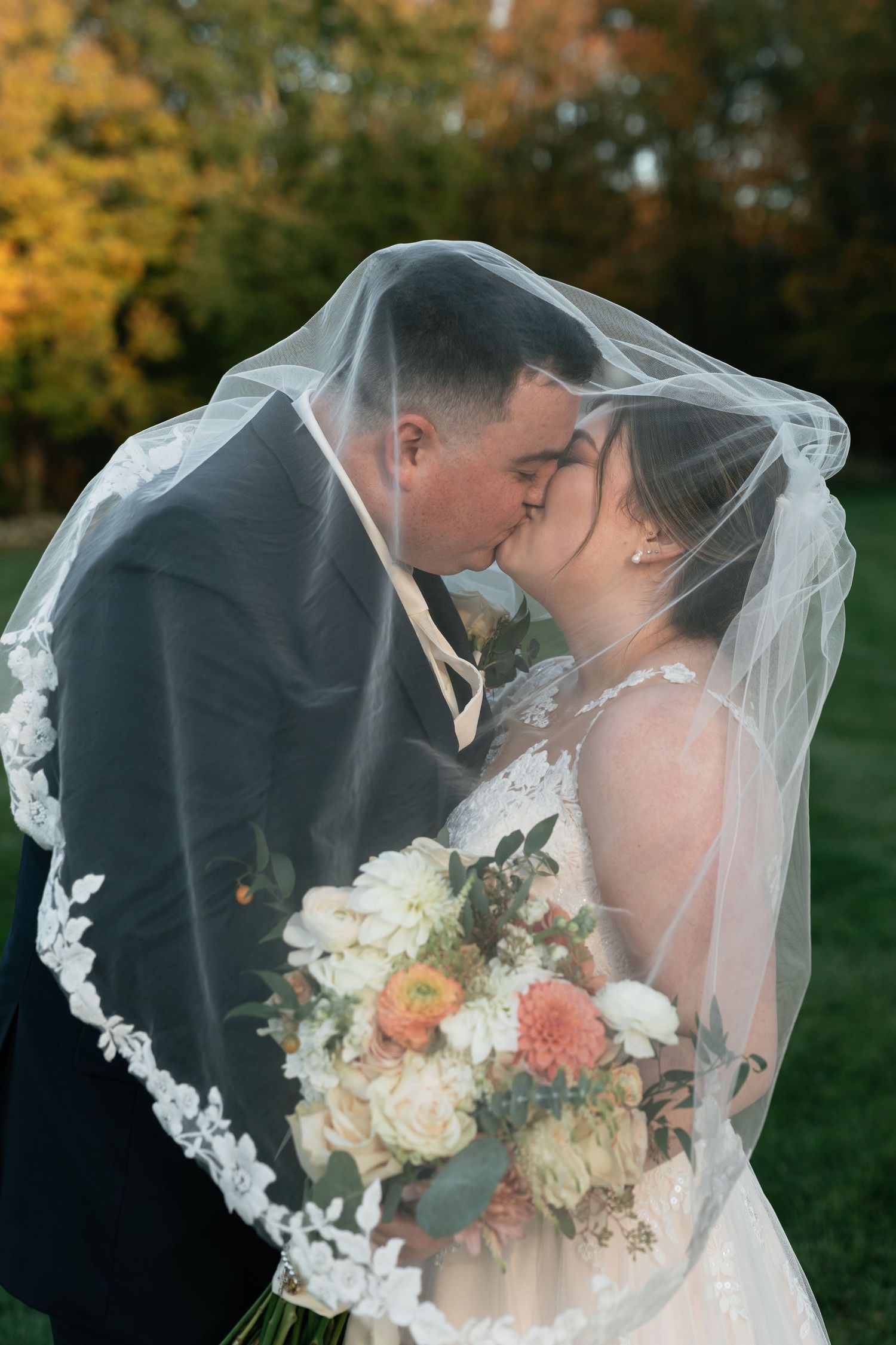 Romantic wedding moment under lace veil with autumn foliage in background.