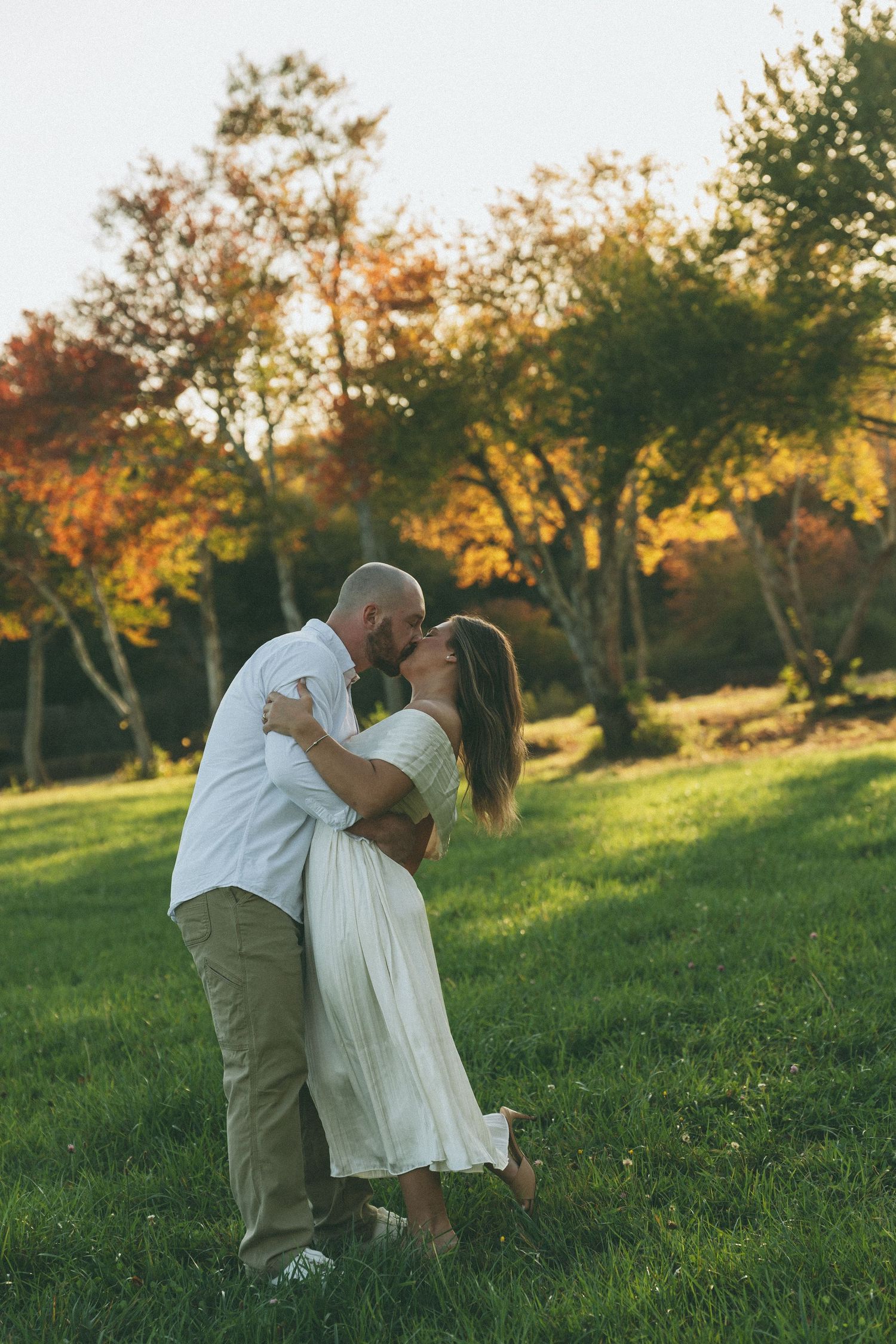 A couple in white and khaki dances in a grassy field at sunset.