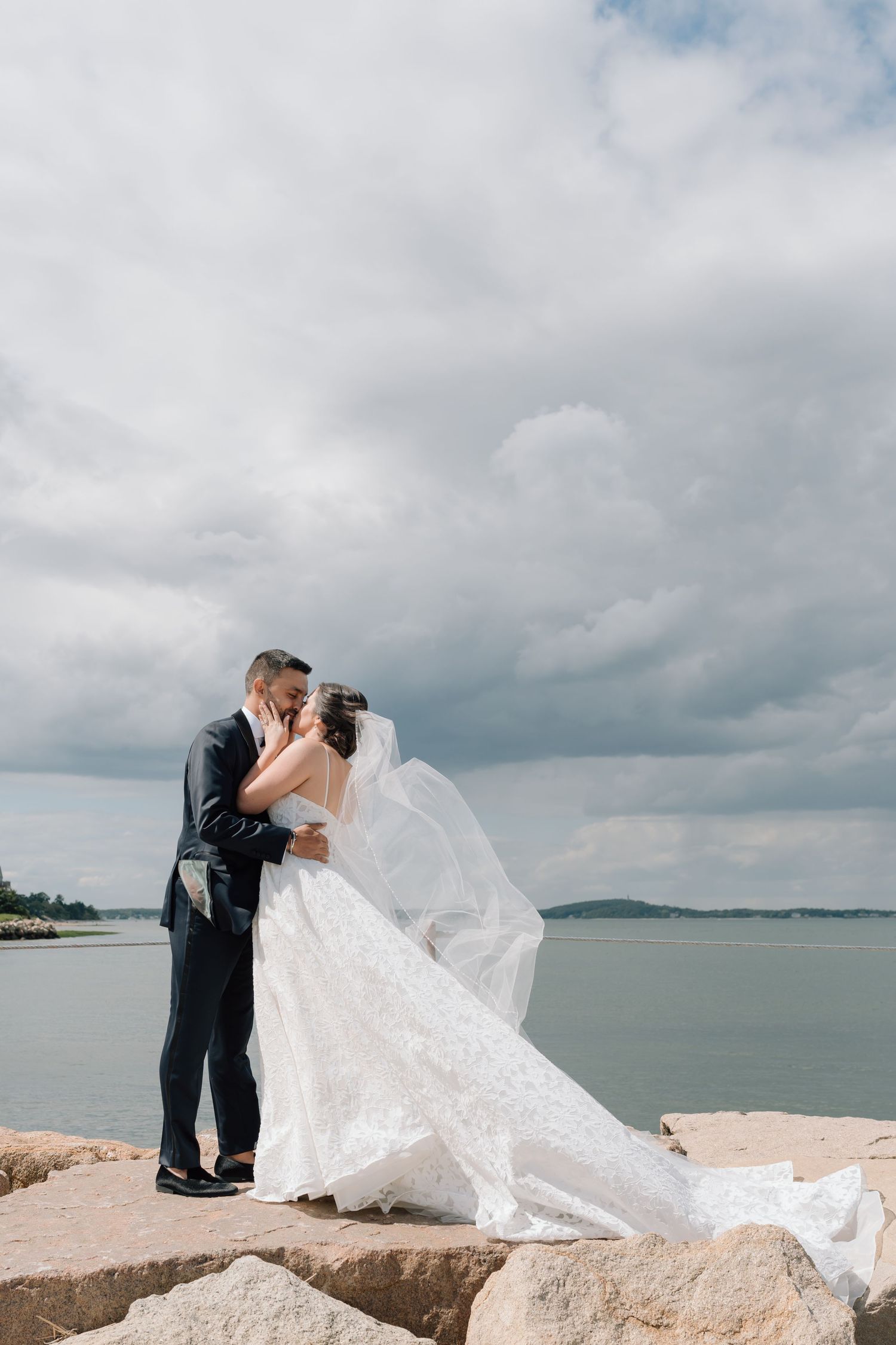 Romantic wedding couple embracing on rocky shoreline with flowing veil and dramatic cloudy sky.