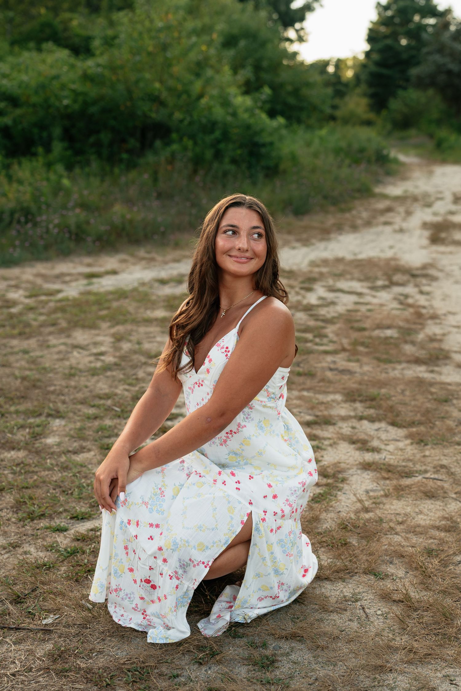 Person in floral white sundress poses on dirt path during outdoor photoshoot.