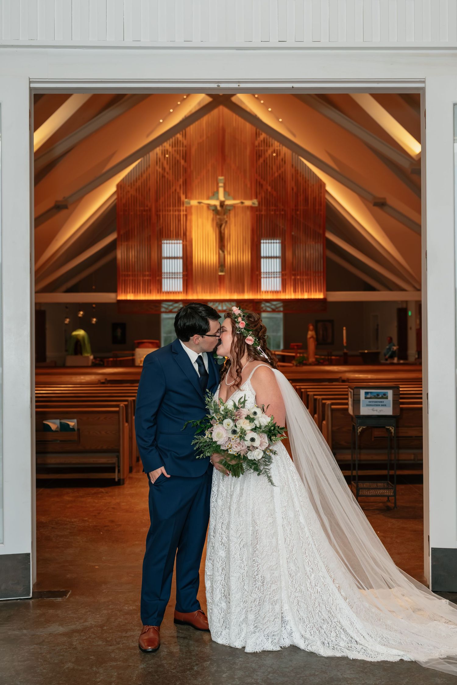 Newlyweds share intimate moment in church doorway with wooden crucifix visible behind.