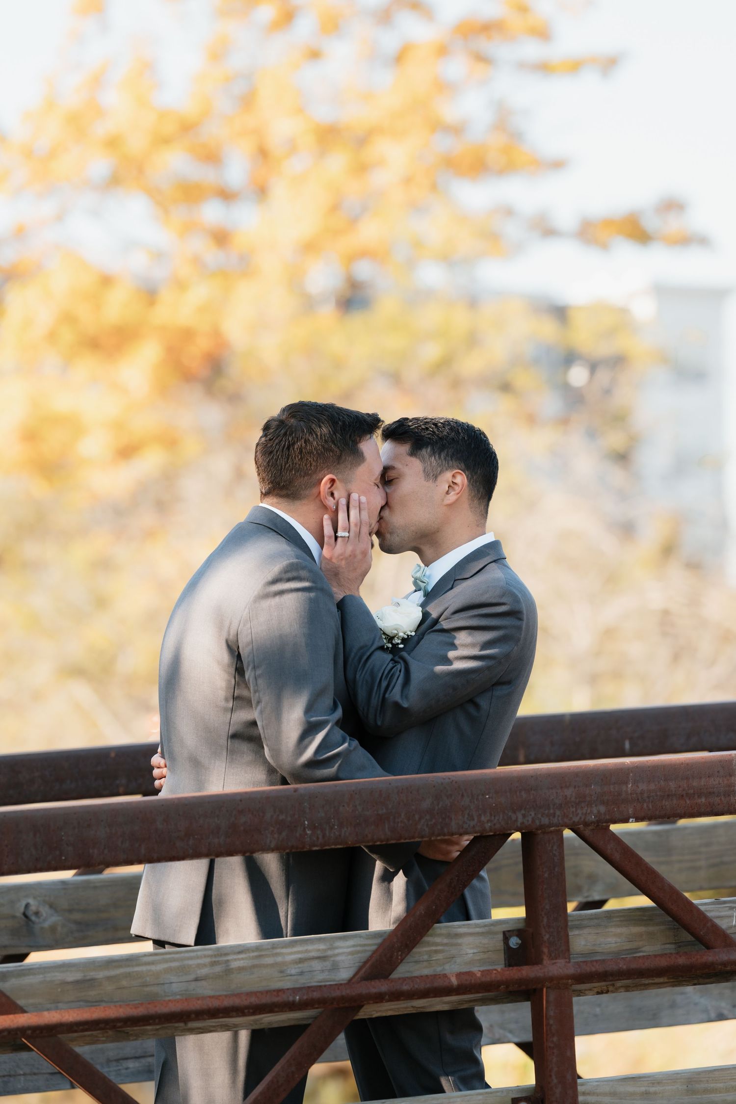 Couple shares an intimate moment on a wooden bridge with fall foliage in background.