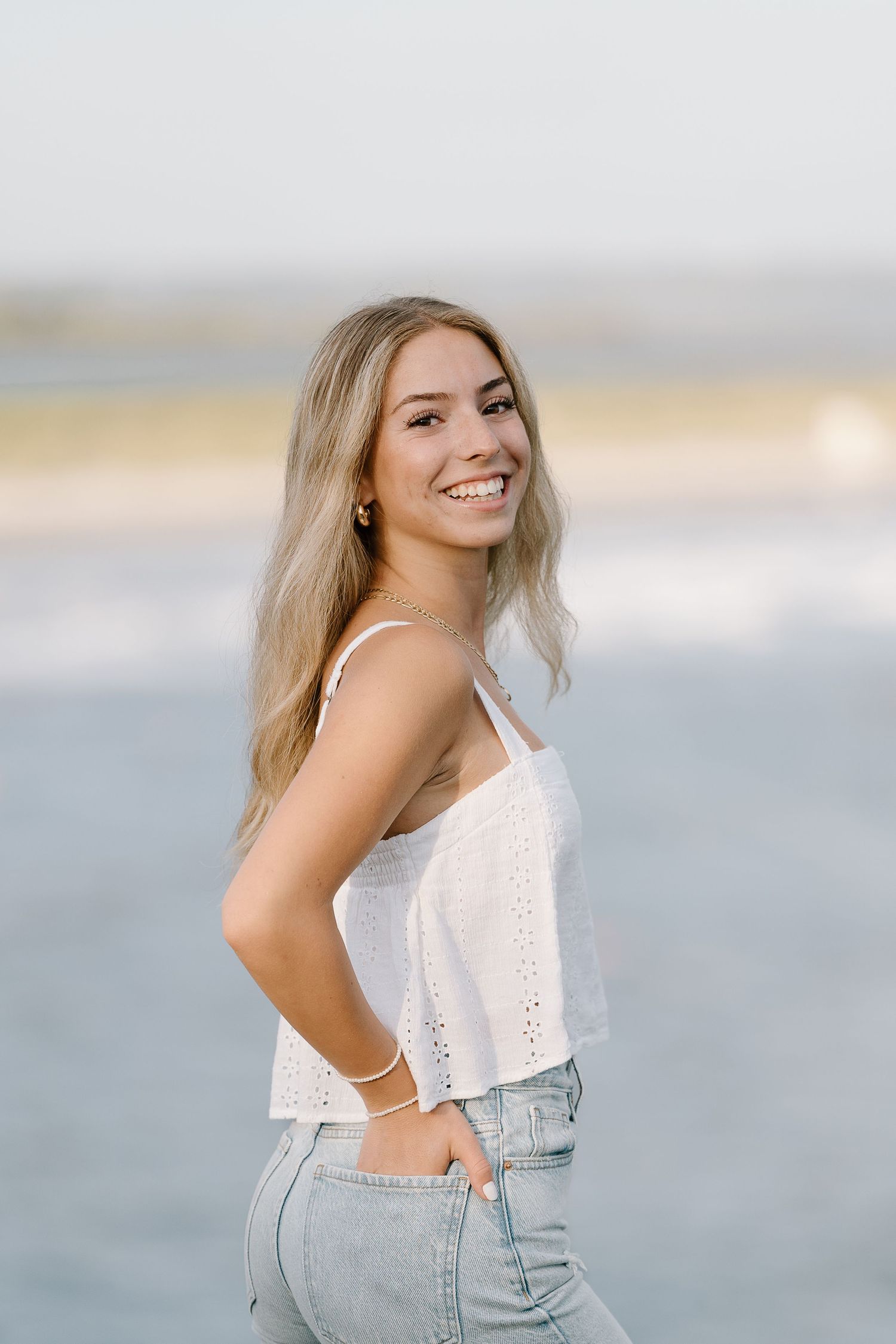 Casual pose in white tank top and jeans with ocean view behind.