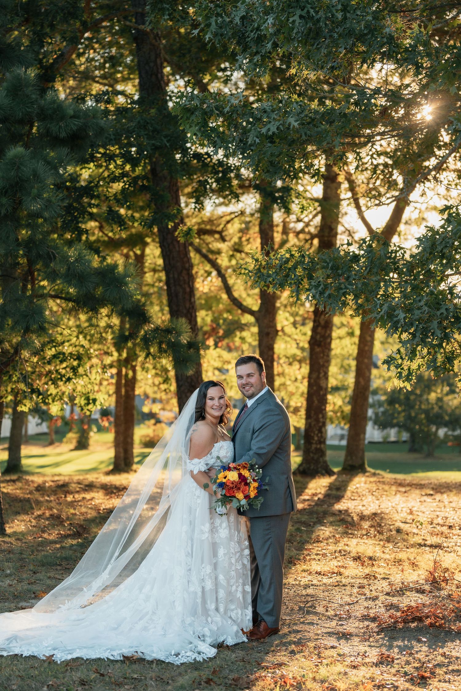 Wedding couple poses in golden sunlight filtering through trees in an outdoor setting.