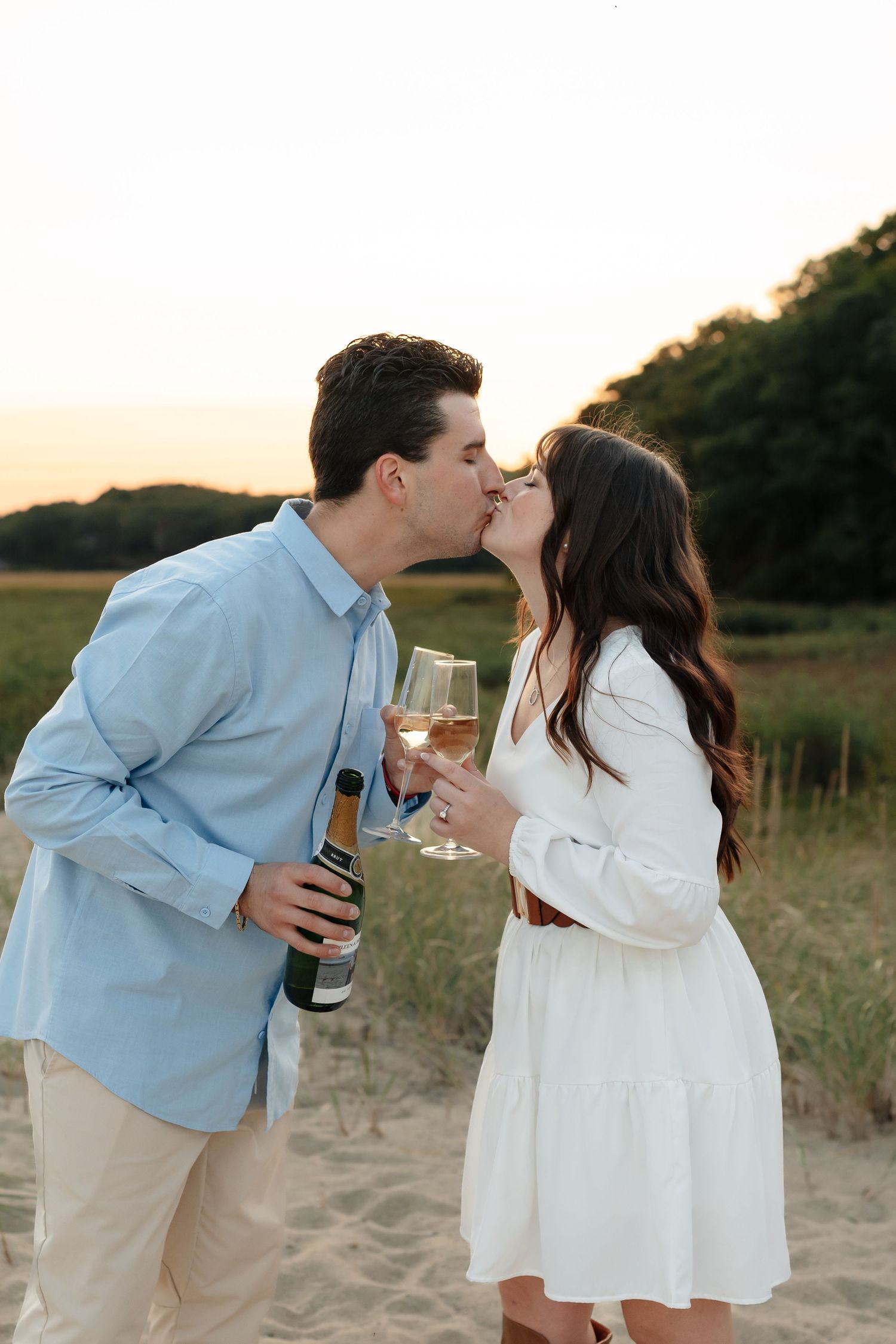 A couple shares drinks and kisses on a sandy beach at sunset.