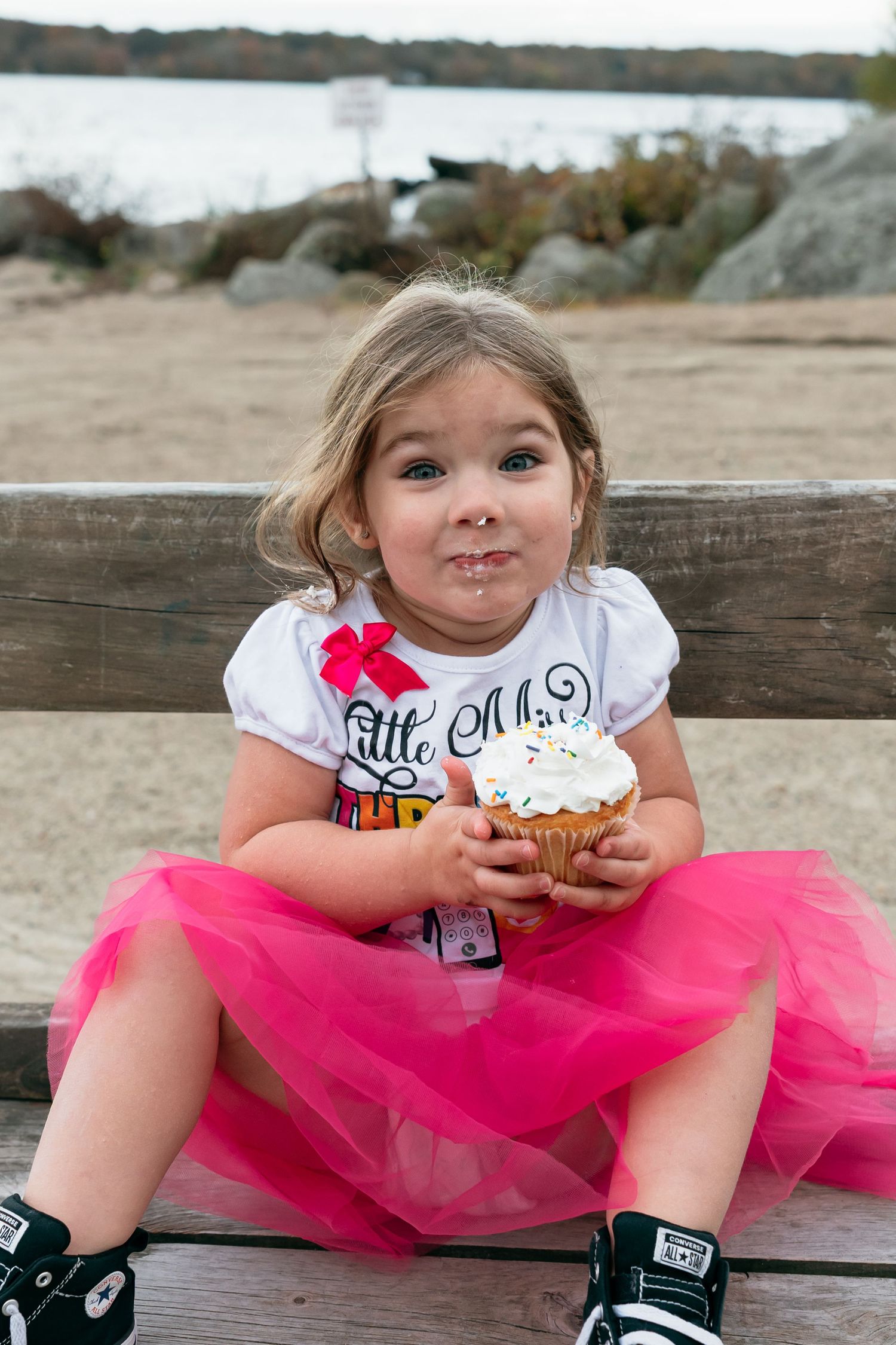 A child in a bright pink tutu and white t-shirt sits on a bench enjoying a cupcake at the beach.