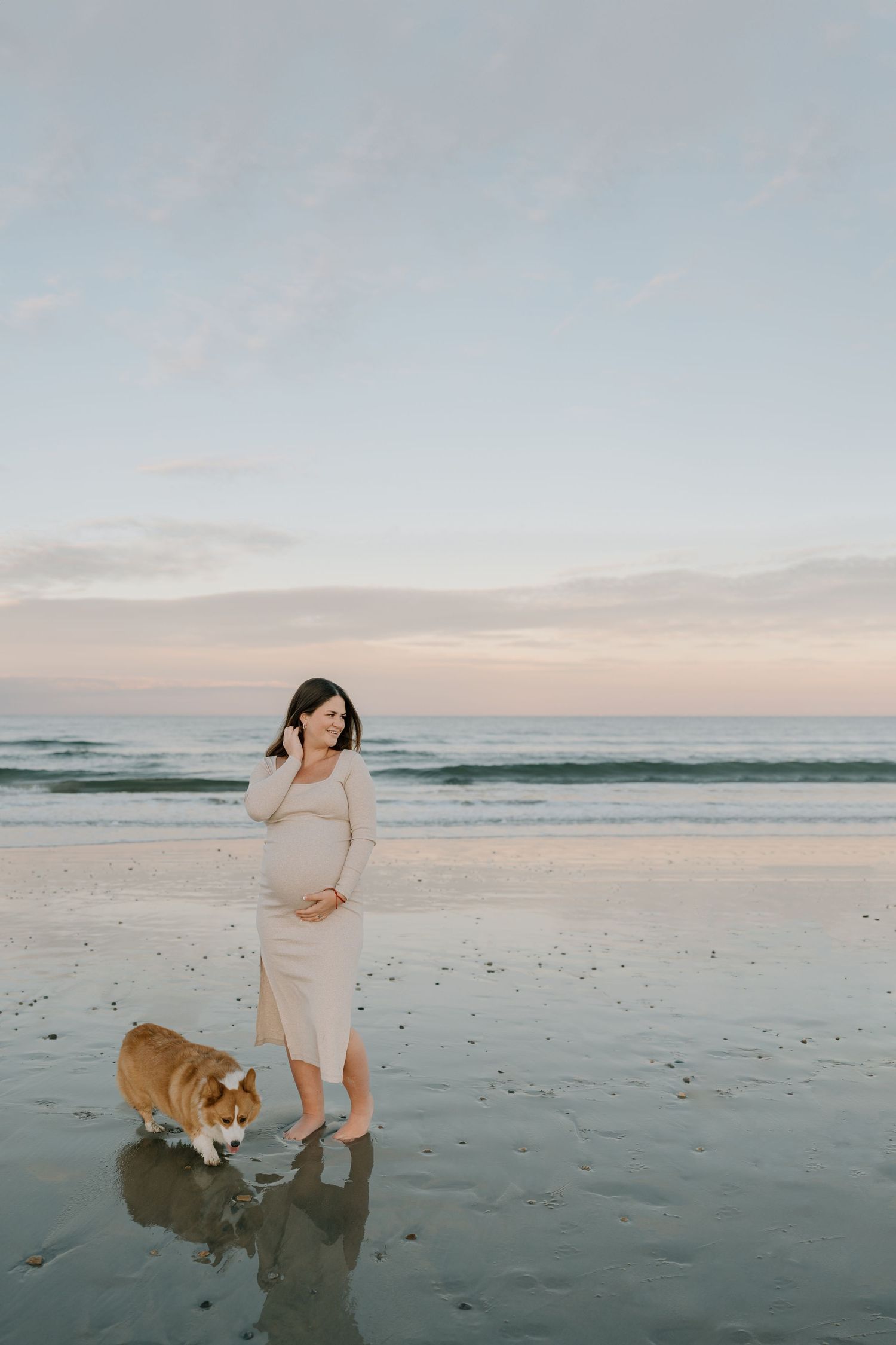 A person in a light dress walks along the beach with a dog at sunset.