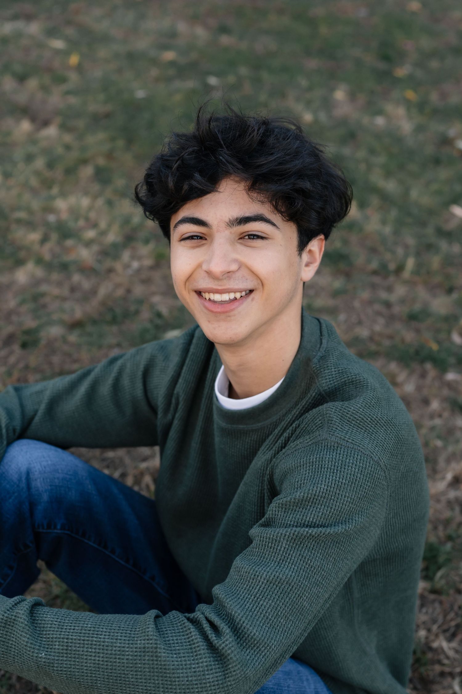 Person in green sweater and jeans sitting outdoors with a cheerful expression.